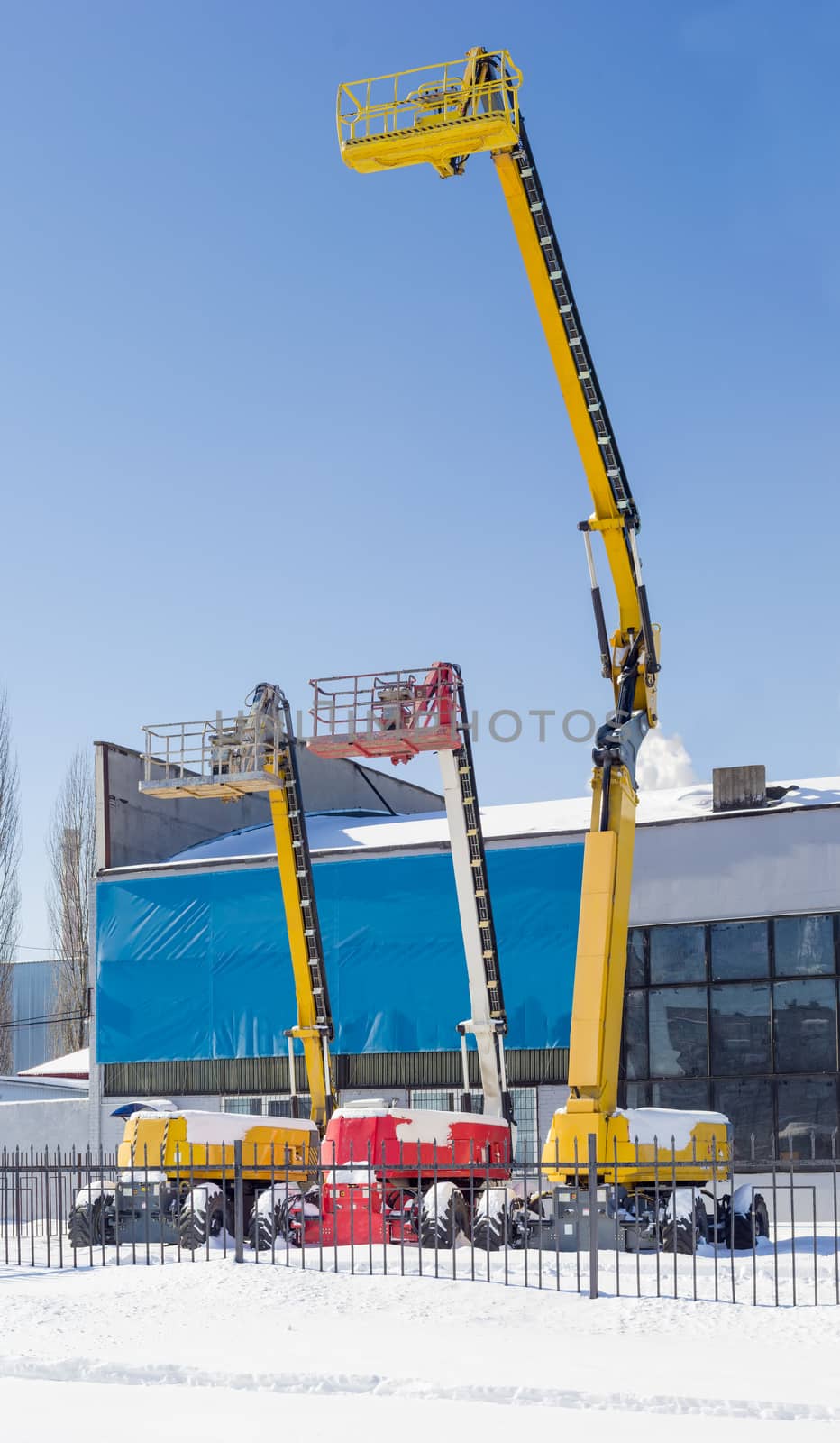 Vertical panorama of the three different self propelled wheeled hydraulic articulated boom lift with telescoping booms and baskets against the sky and an industrial building in winter sunny day
