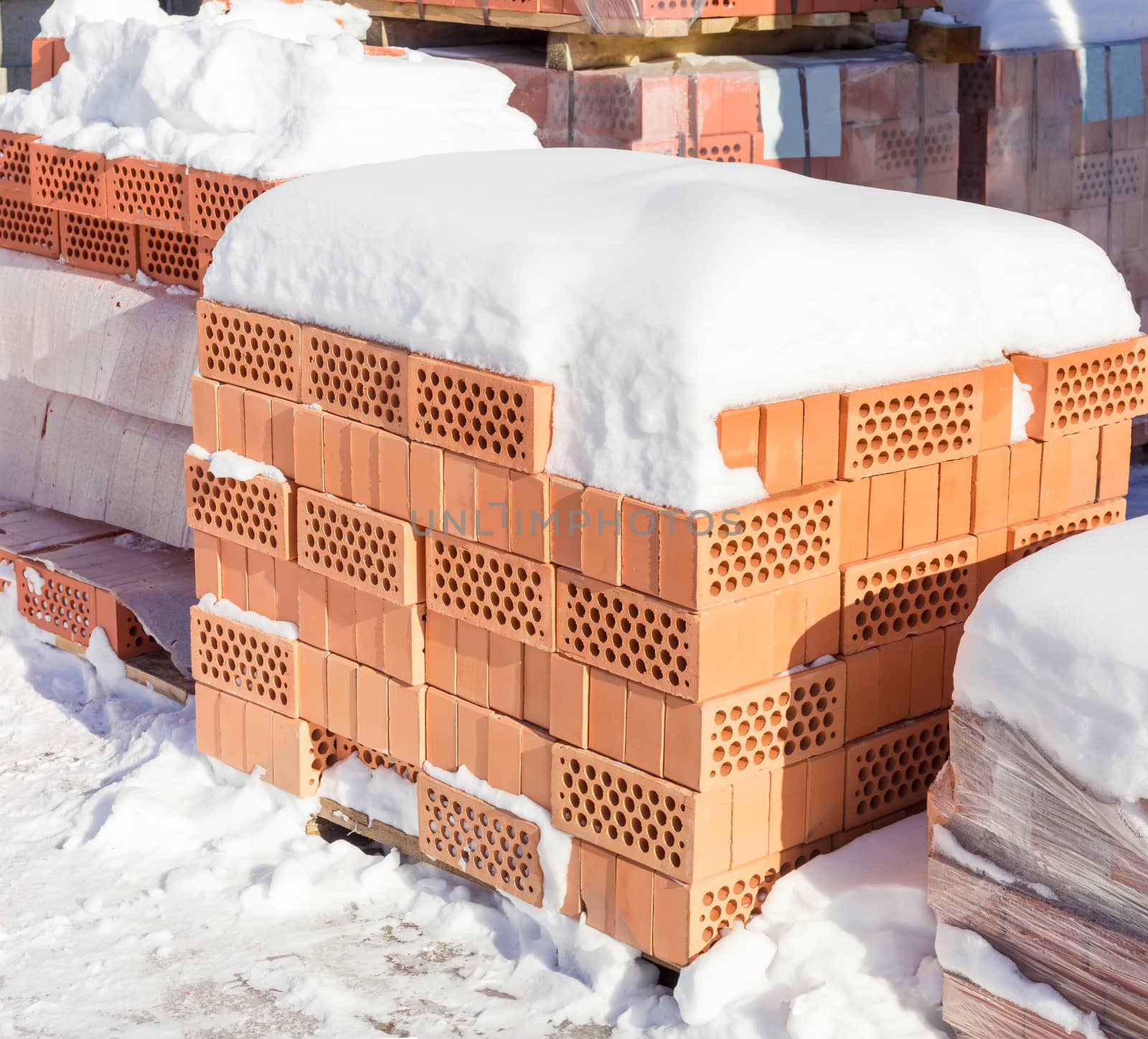 Red perforated bricks with round holes covered snow on a pallet against the background of other pallets with bricks on an outdoor warehouse in winter sunny day
