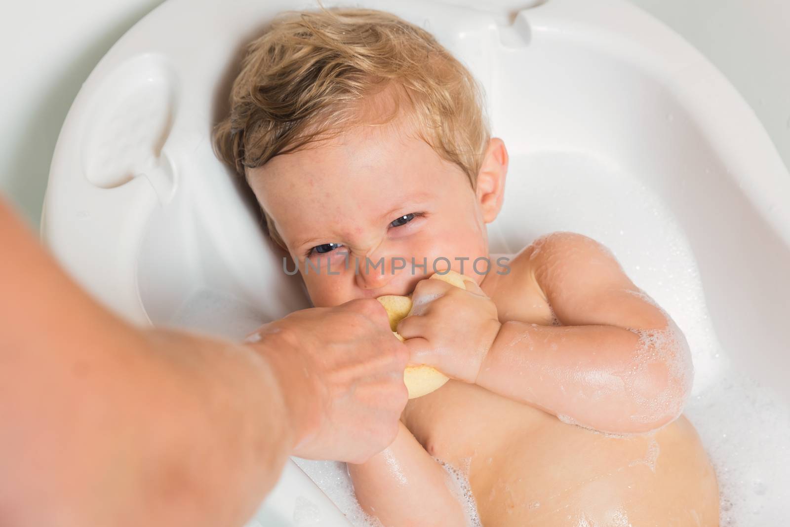 Little pretty wet baby boy in bath room sitting and playing with mother's hand and sponge on white background, horizontal picture.