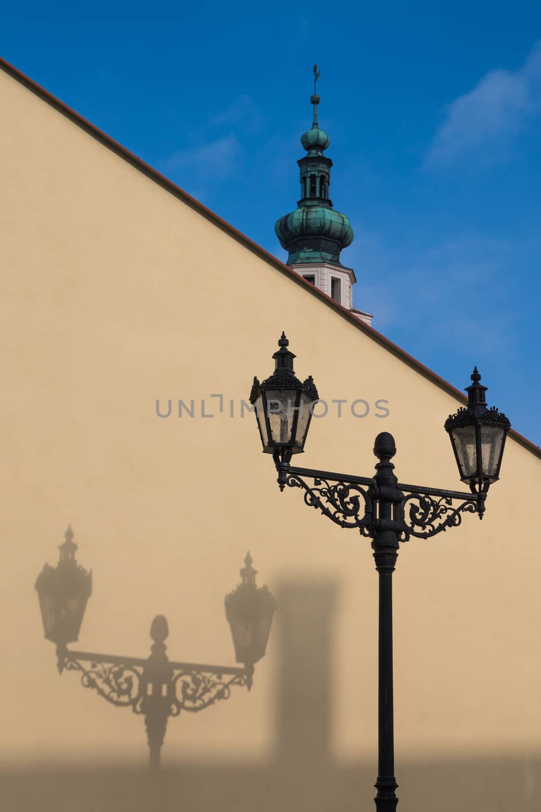 Traditional lantern with its shadow on the yellow diagonal wall. In the background tower of a church. Boskovice, Czech republic.