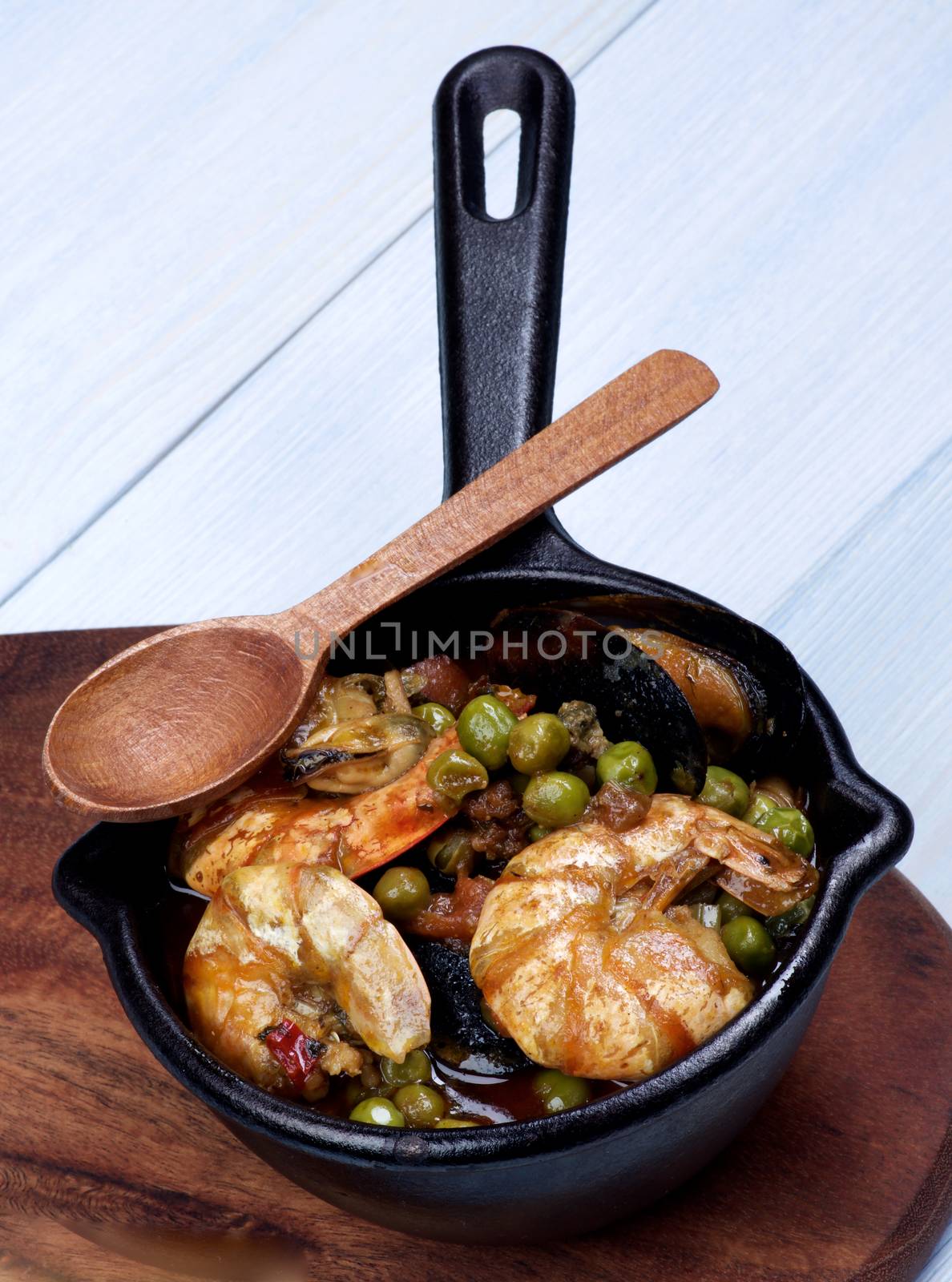 Delicious Seafood Curry with Prawns, Mussels, White Fish and Vegetables in Black Iron Cast Pot with Wooden Spoon closeup on Light Blue background