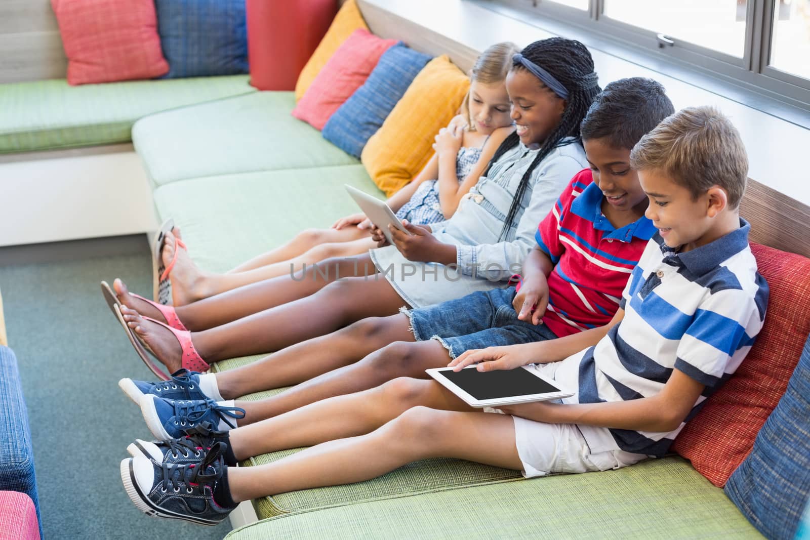 School kids sitting on sofa and using digital tablet in library by Wavebreakmedia
