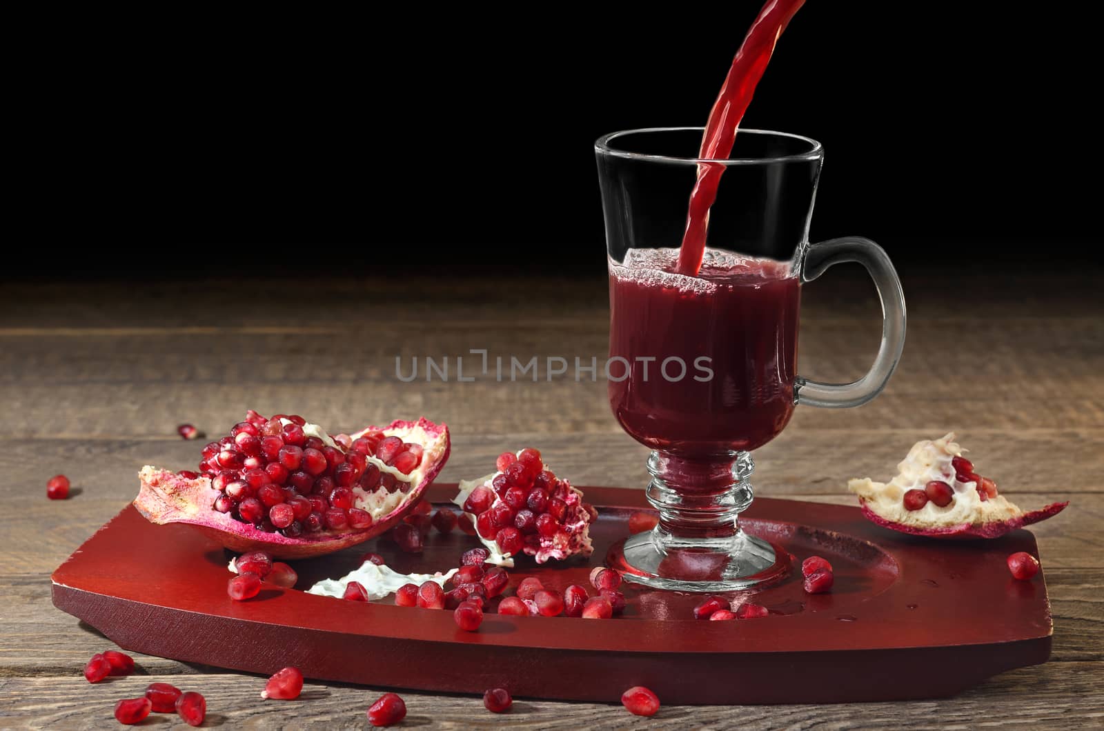 Pomegranate juice is poured into a glass Cup, wooden surface and black background. Pieces and pomegranate seeds on a tray. Selective focus.