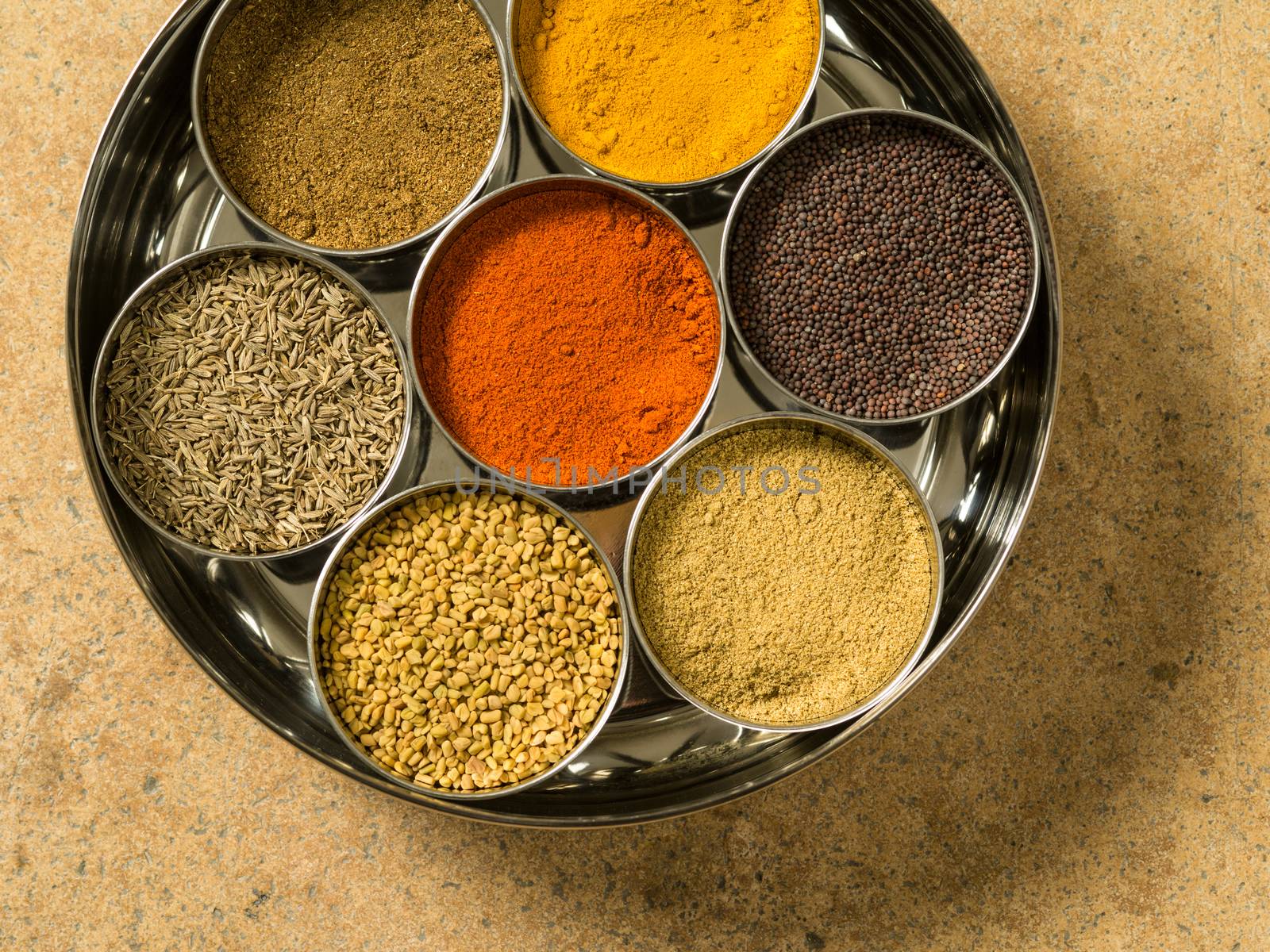 Photo of a Masala box containing turmeric, fenugreek, coriander, garam masala, black mustard seeds, red chilli, and cumin seeds.