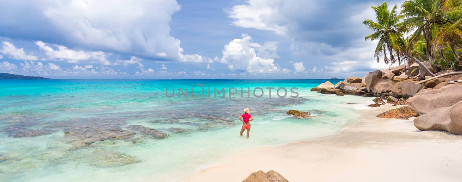 Woman wearing retro striped bikini and beach hat, enjoying amazing view on Anse Patates beach on La Digue Island, Seychelles. Summer vacations on picture perfect tropical beach concept.