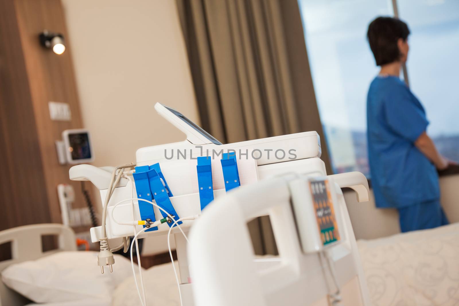 Cardiac stress test equipment in a modern hospital room with a blurred figure of a patient looking through the window.