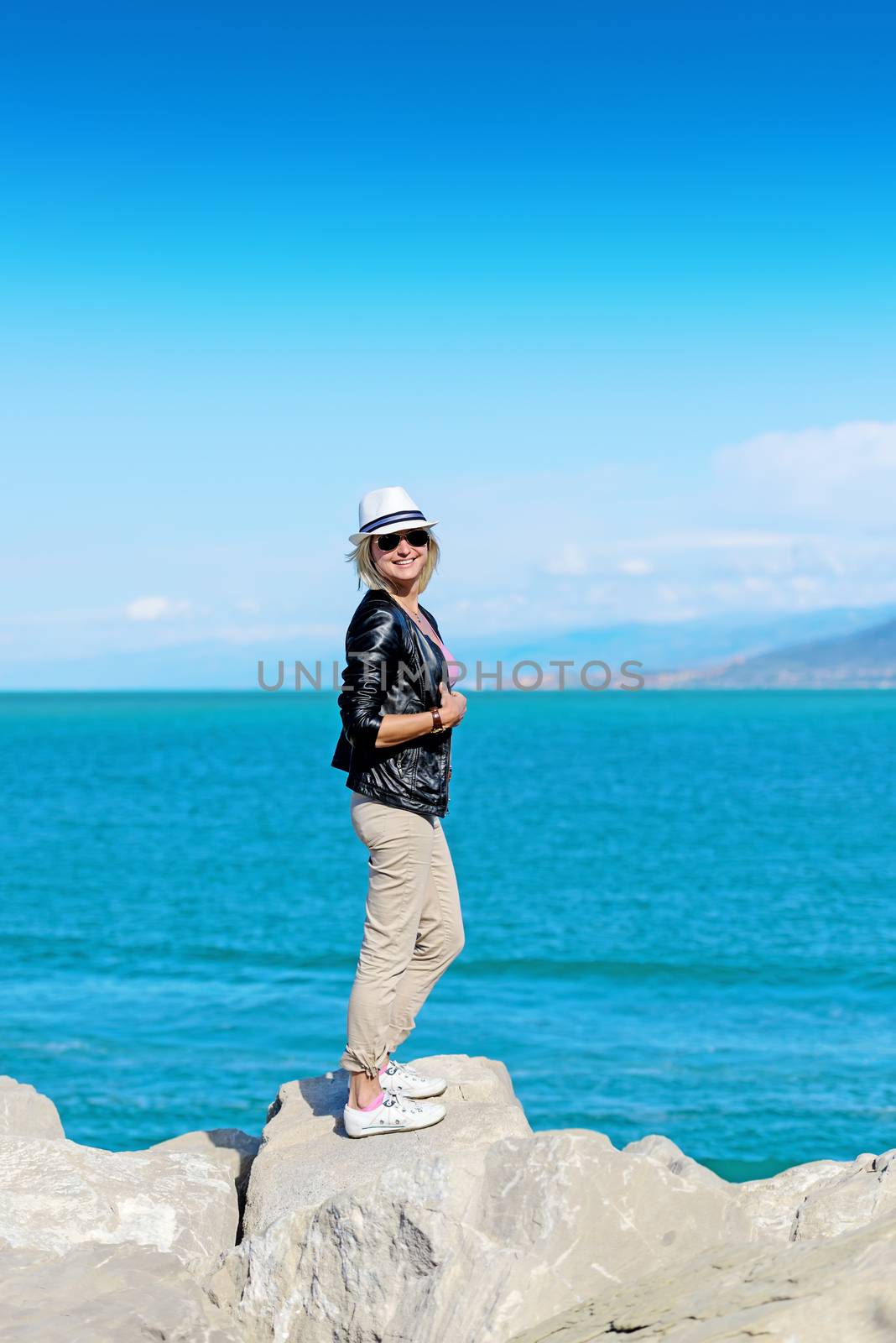 Smiling blond woman in hat and sun glasses standing on a rock on coastline
