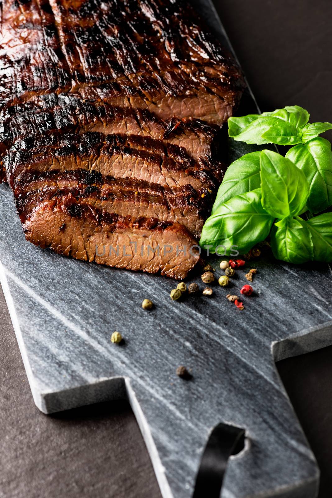 Close up sliced medium grilled beef steak with pepper and oregano leafs on marble meat cutting board on dark stone background