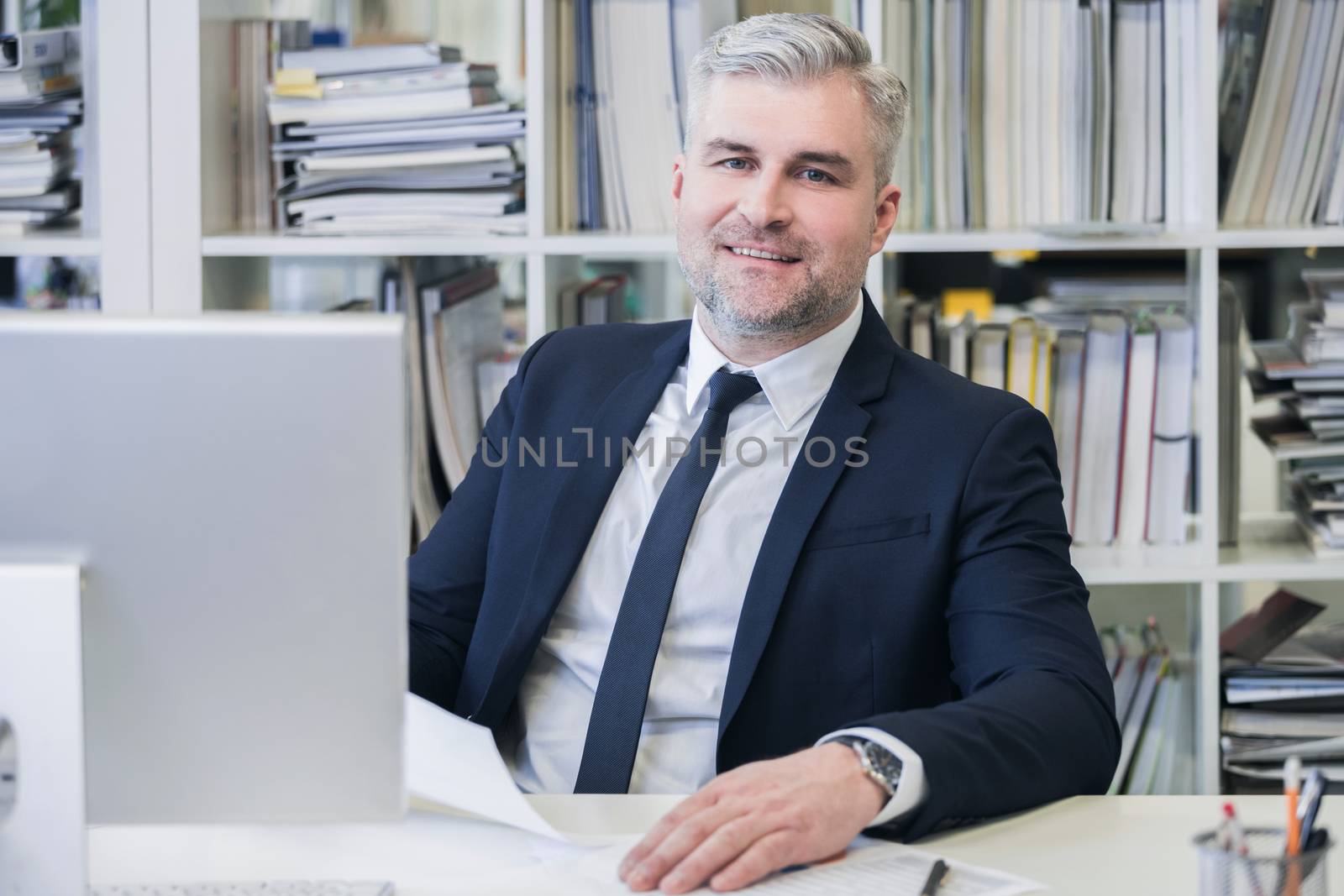 Portrait of mature business man with grey hair in his modern office with computer and many file folders on shelves on background