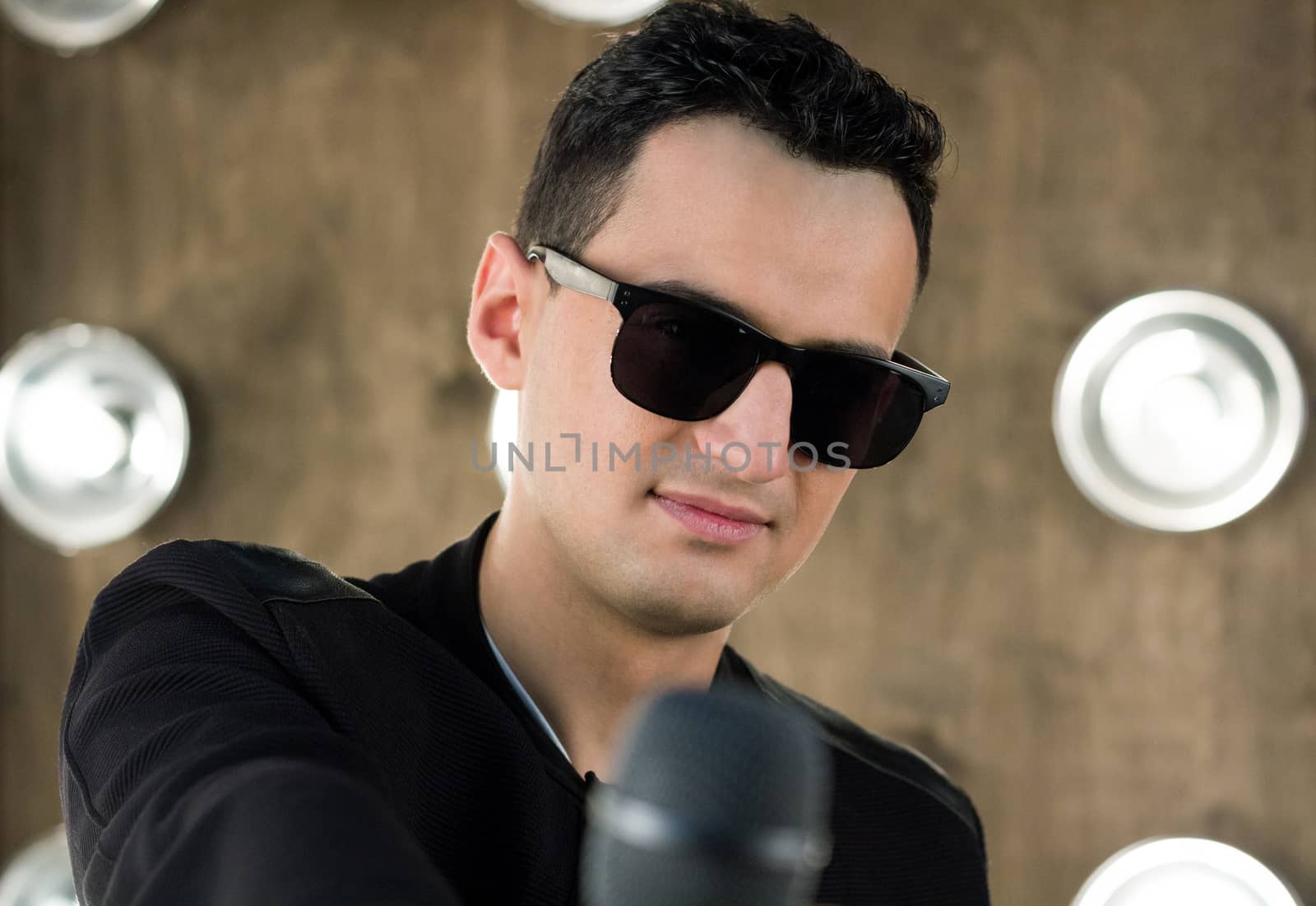 Male singer of rock or pop music dressed in black and sunglasses with microphone performs on scene with lightening projectors on background