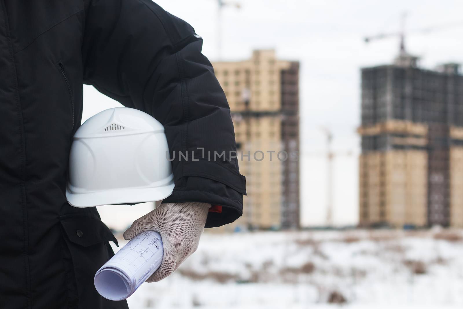 Architect or engineer holding yellow helmet for workers security on background of new highrise apartment buildings and construction cranes on background of winter landscape. Crane lifts load