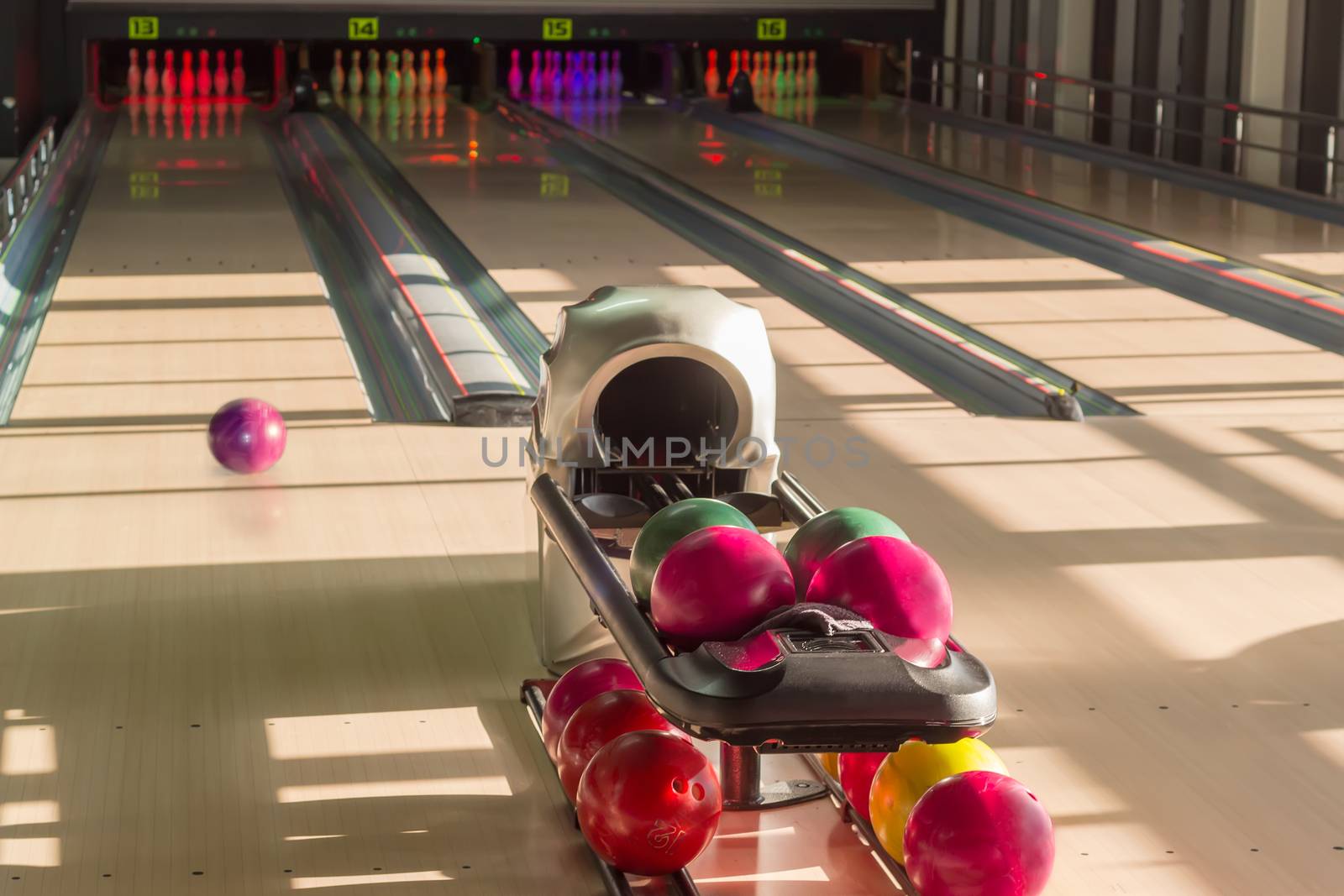 Playing area with several lanes with bowling pins, bowling ball rolling along the lane and colored bowling balls on the foreground in the modern pin bowling alley

