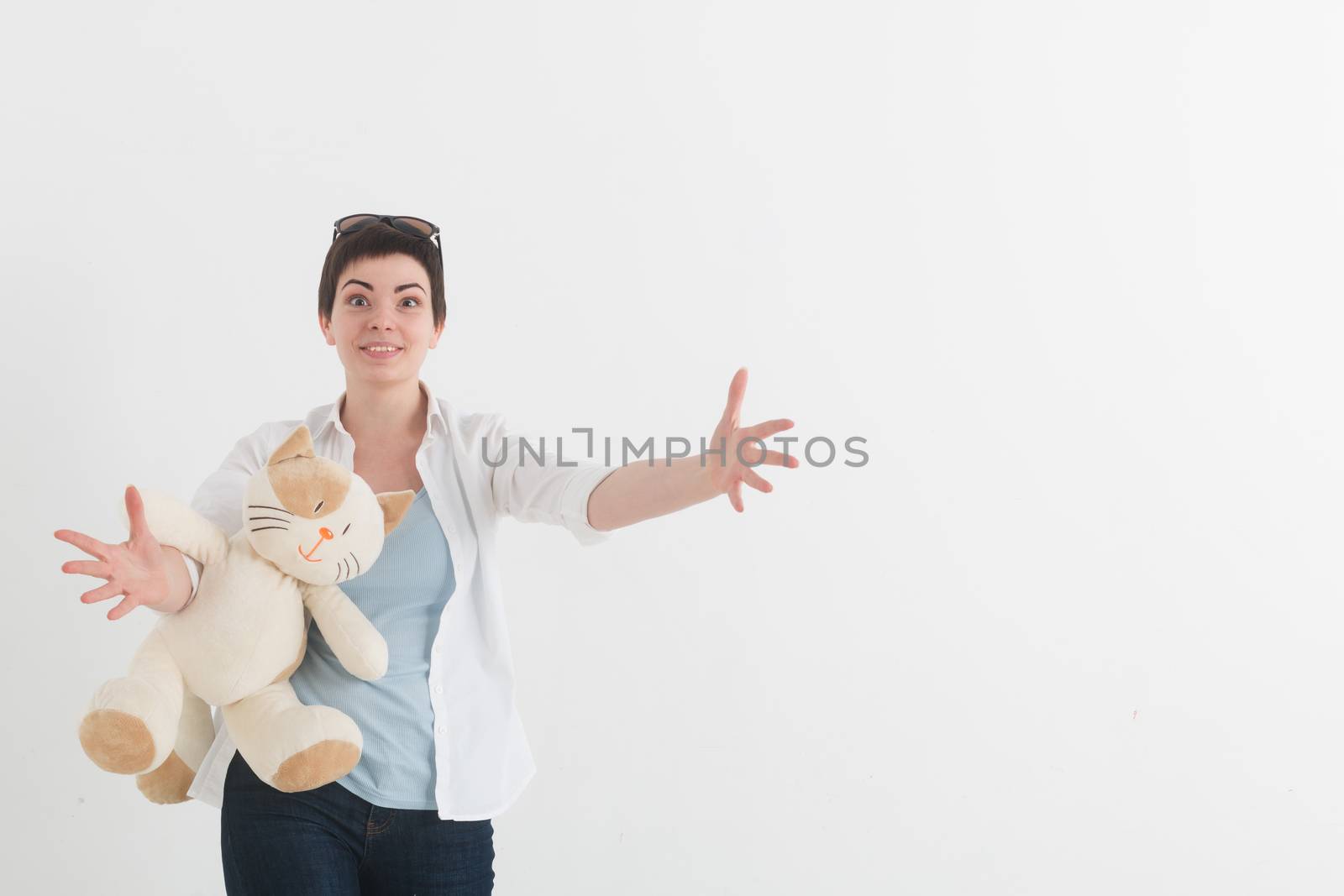 Portrait of a young girl in white shirt on the light background. Looking at the camera, smiling and extending hands forward to the spectator. Happy woman with plush toy cat.