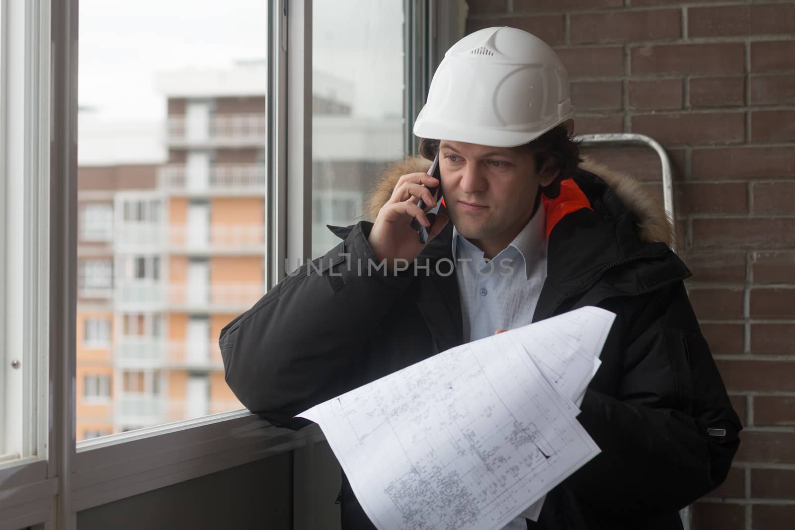 Engineer standing on the background of a new apartment building. House is built. Engineer in a helmet with papers in hand on the phone. The concept of new housing construction, real estate.