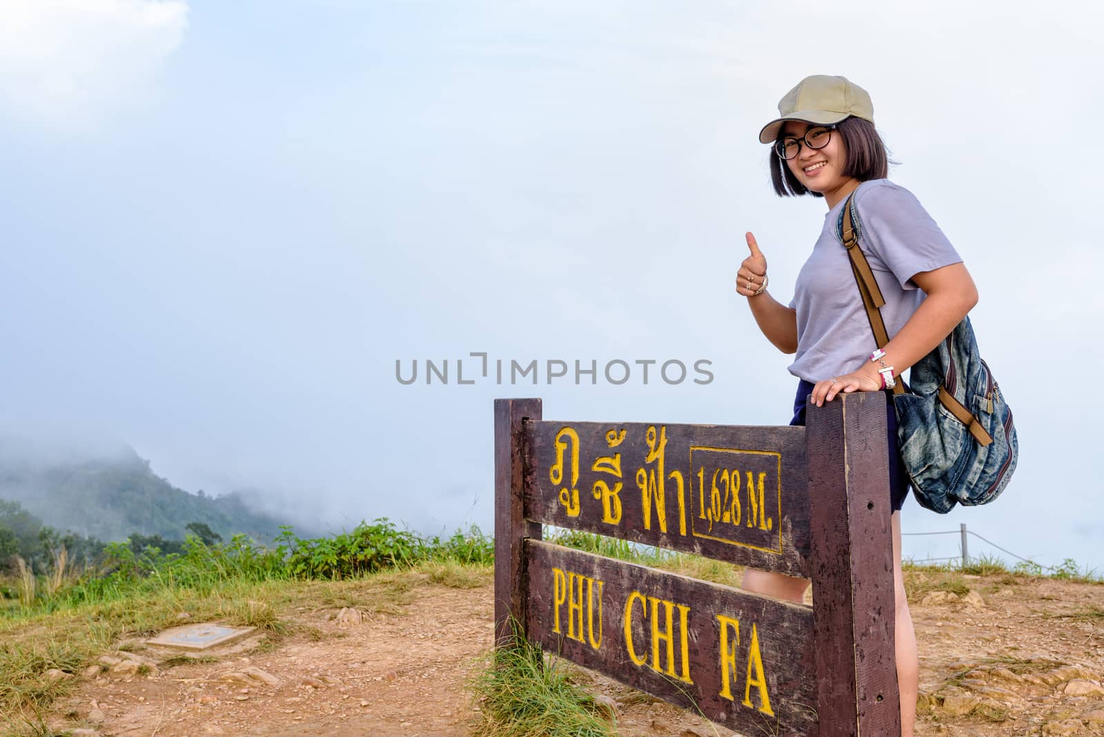 Tourist teen girl hiker wear cap and glasses with backpack smiling thumb up near nameplate attractions of Phu Chi Fa Forest Park on mountain and mist background, Chiang Rai, Thailand