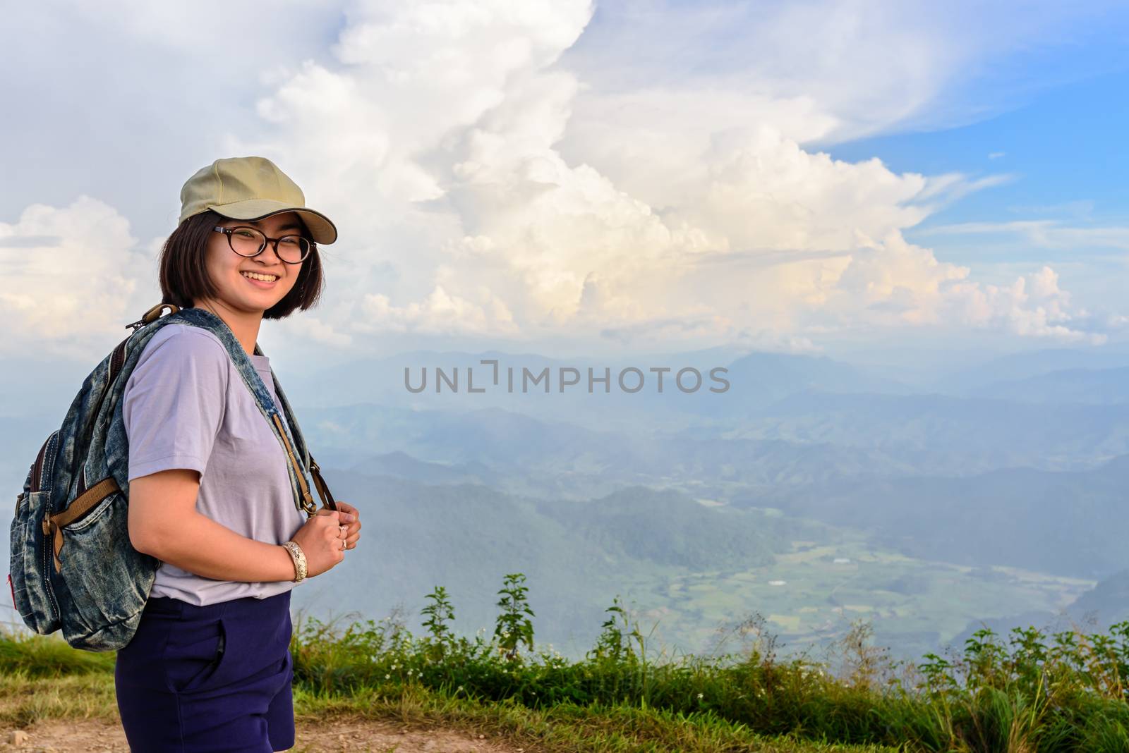 Teens girl hiker wear a cap and glasses with backpack is standing and smiling happily on high mountain at scenic point of Phu Chi Fa Forest Park in Chiang Rai Province Thailand