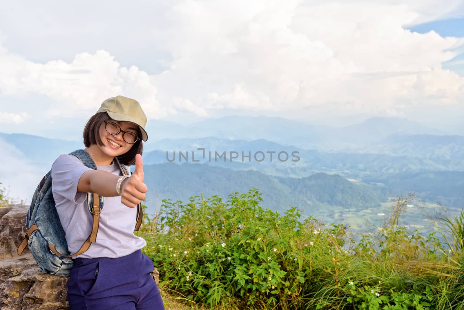 Tourist teens girl hiker wear cap and glasses with backpack poses thumb up smiling happily on high mountain at scenic point of Phu Chi Fa Forest Park in Chiang Rai Province Thailand