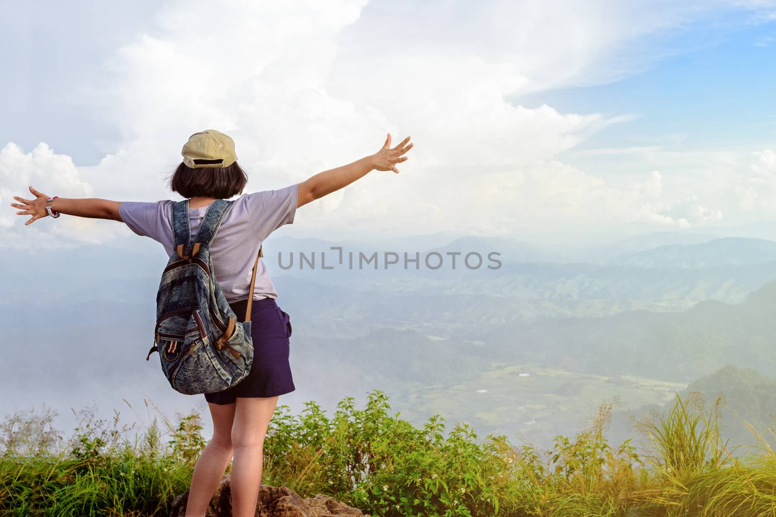 Happy teens girl hiker with backpack and cap open arms looking beautiful nature landscape on high mountain at viewpoint of Phu Chi Fa Forest Park during sunset in Chiang Rai Province Thailand