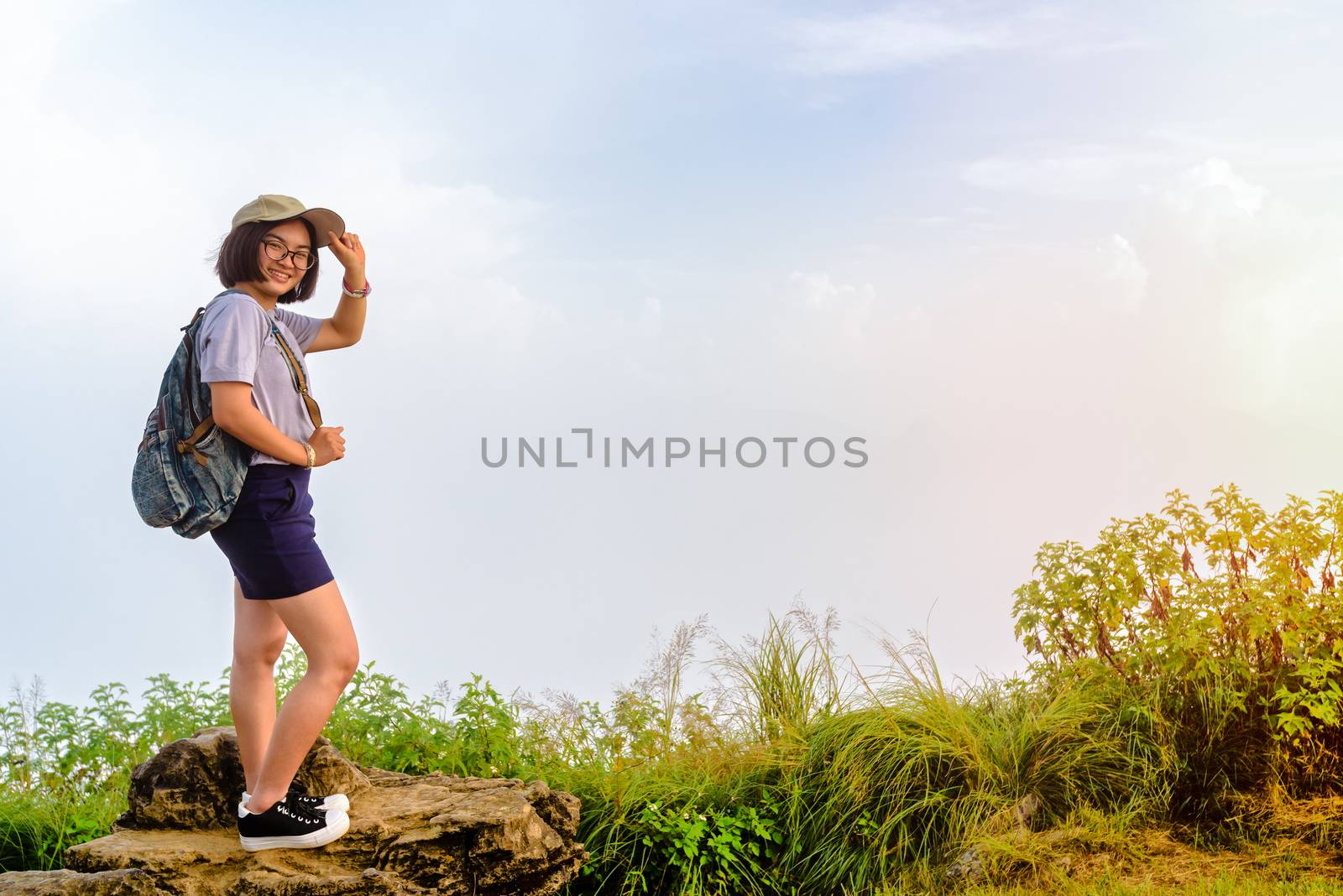 Tourist teens girl hiker with backpack cap and glasses is standing smile and poses happily at high mountain on sky and fog background at scenic point of Phu Chi Fa Forest Park in Chiang Rai, Thailand