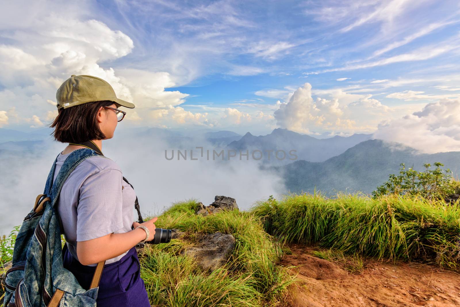 Hiker asian teens girl with camera and backpack wear caps and glasses looking beautiful landscape nature of mountain range and blue sky during sunset at Phu Chi Fa Forest Park, Chiang Rai, Thailand