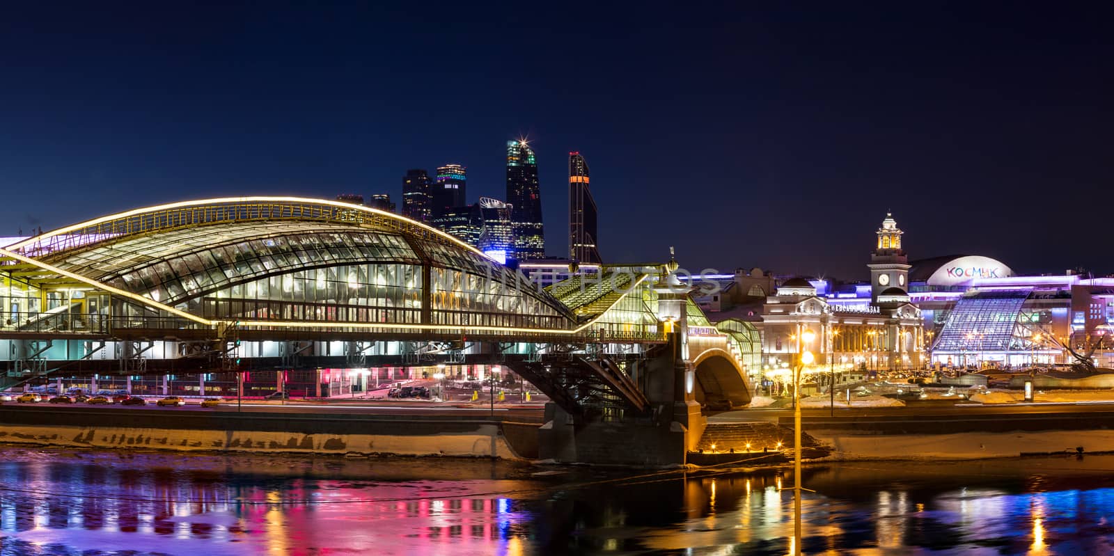 Night winter panorama of the Moskva River embankment: pedestrian bridge Bogdan Khmelnitsky, Kiyevskaya railway station, Moscow-City, Redisson Slavyanskaya hotel, "Europe" Square, shopping center "European". Rich colorful illuminations and reflections in the river.