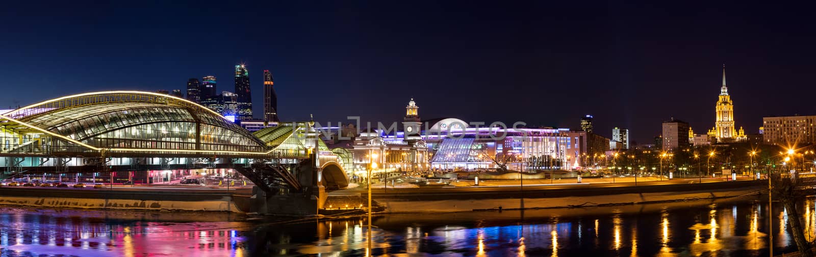 Night winter panorama of the Moskva River embankment: pedestrian bridge Bogdan Khmelnitsky, Kiyevskaya railway station, Moscow-City, Redisson Slavyanskaya hotel, "Europe" Square, shopping center "European", Radisson Royal Hotel (the former "Ukraine"). Rich colorful illuminations and reflections in the river.