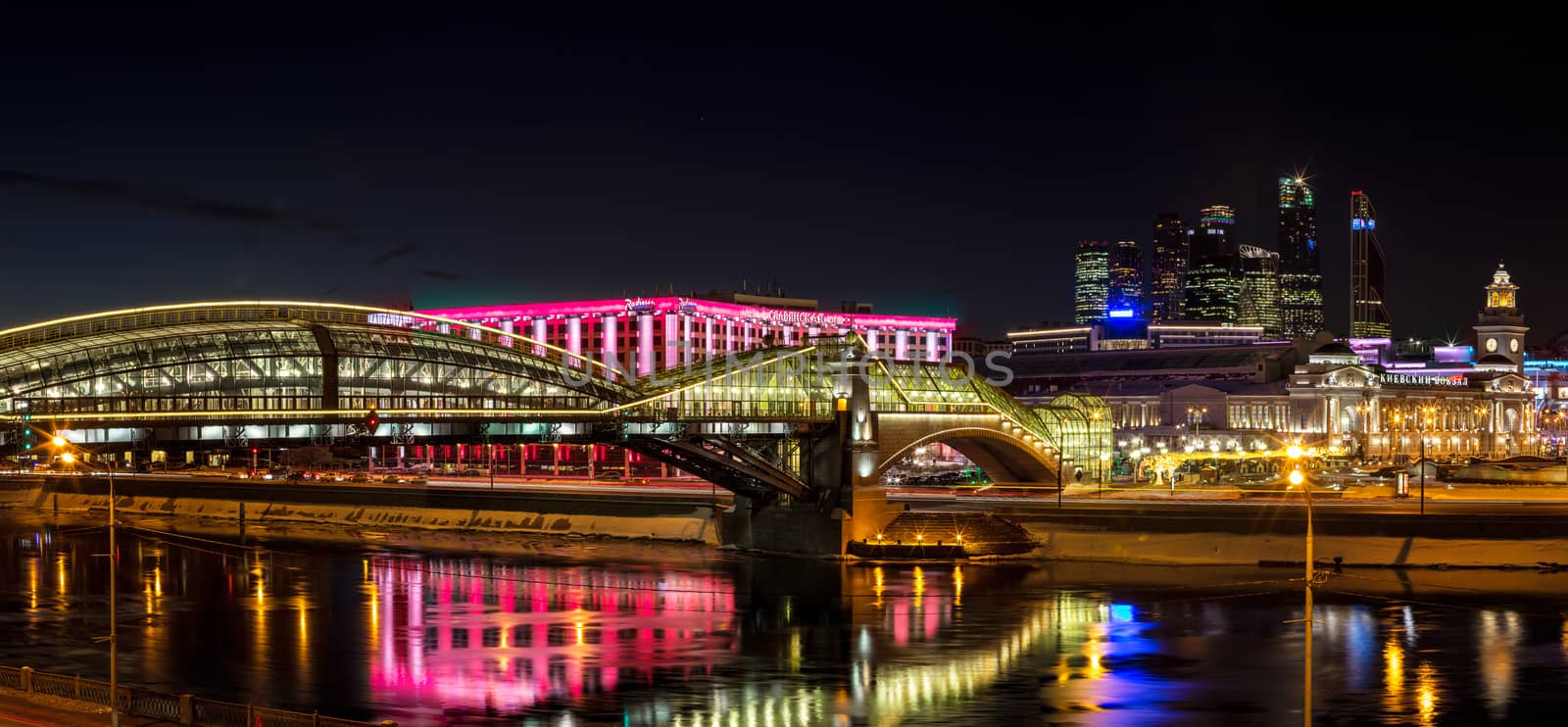 Night winter panorama of the Moskva River embankment: pedestrian bridge Bogdan Khmelnitsky, Kiyevskaya railway station, Moscow-City, Redisson Slavyanskaya hotel. Rich colorful illuminations and reflections in the river.