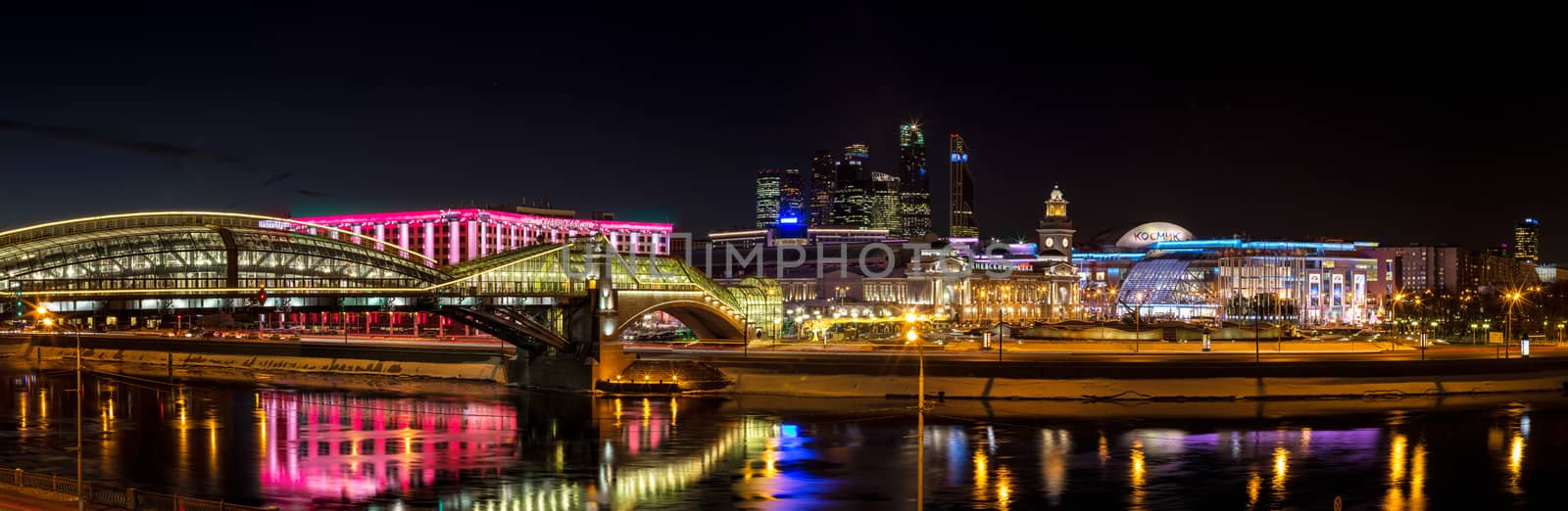 Night winter panorama of the Moskva River embankment: pedestrian bridge Bogdan Khmelnitsky, Kiyevskaya railway station, Moscow-City, Redisson Slavyanskaya hotel, "Europe" Square, shopping center "European". Rich colorful illuminations and reflections in the river.