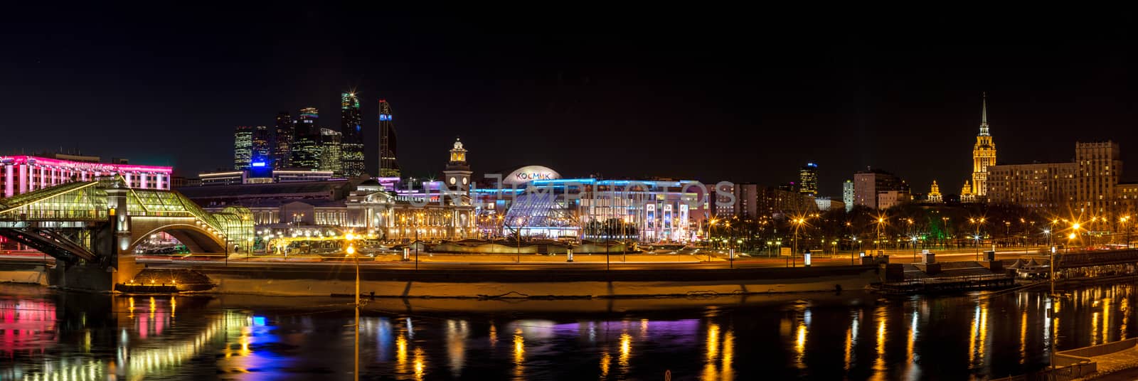 Night winter panorama of the Moskva River embankment: pedestrian bridge Bogdan Khmelnitsky, "Europe" Square, shopping center "European", Kiyevskaya railway station, Moscow-City, Redisson Slavyanskaya hotel, Radisson Royal Hotel (the former "Ukraine"). Rich colorful illuminations and reflections in the river.