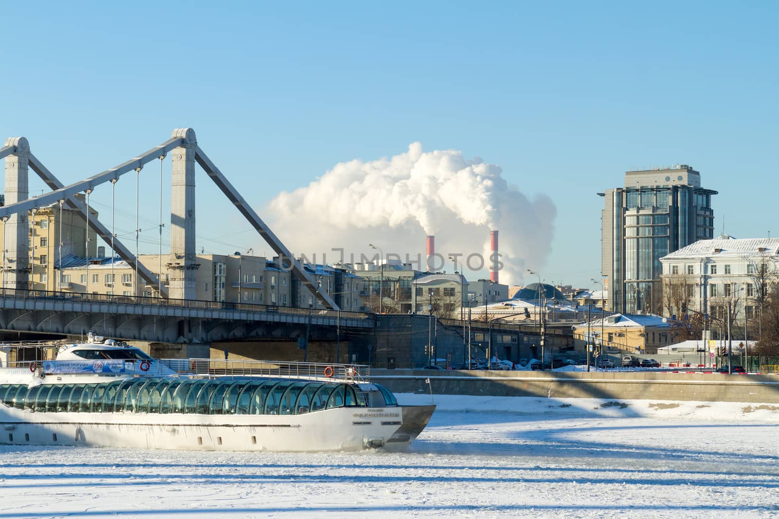 Moscow, Russia - January 8, 2017: Pleasure boat sailing on a frozen river under the bridge metal structures, frosty sunny winter day on the background of residential and office buildings and power plant pipes, spewing steam - in Moscow, Russia