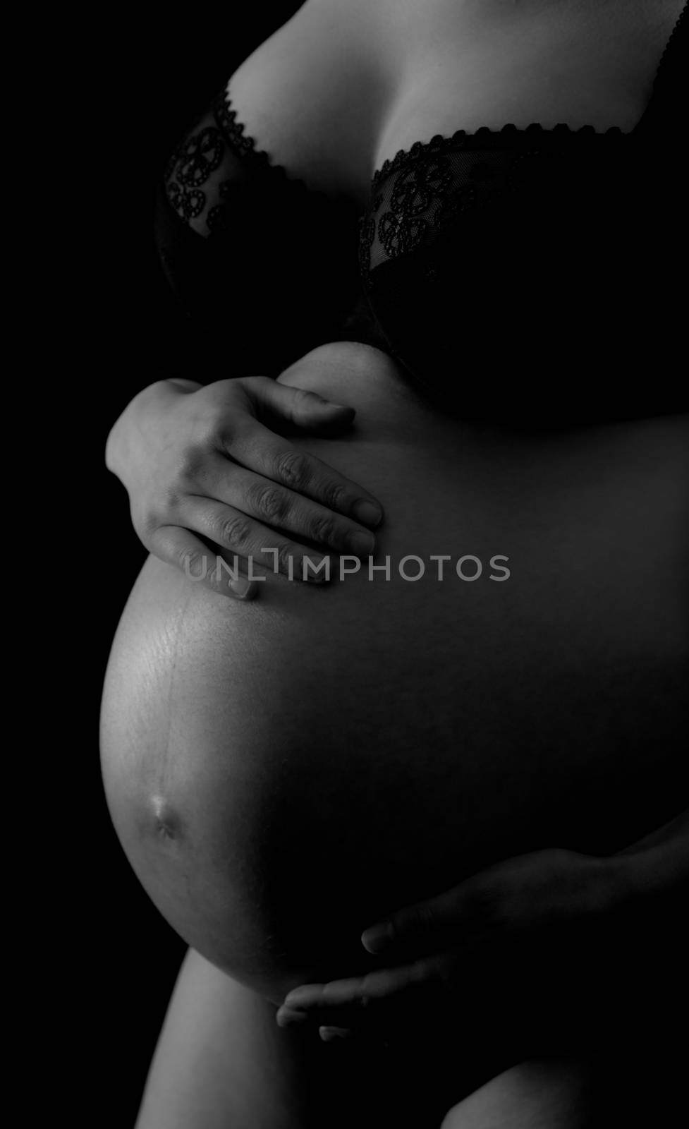 A black and white image of a pregnant woman torso, holding her abdomen with both hands.