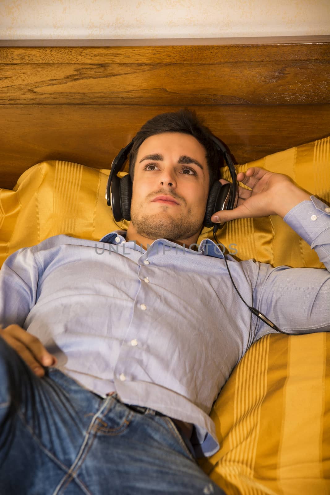 Young man in bed listening to music with headphones by artofphoto