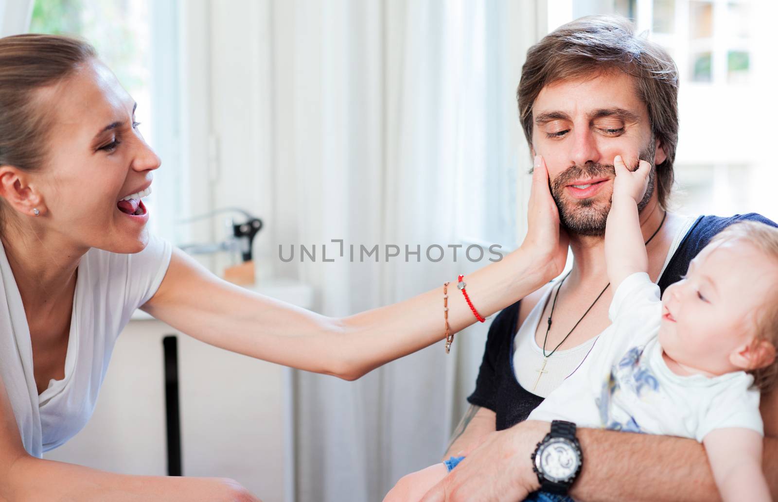 A young mother and a baby are showing affection to their husband and dad pinching his cheeks.