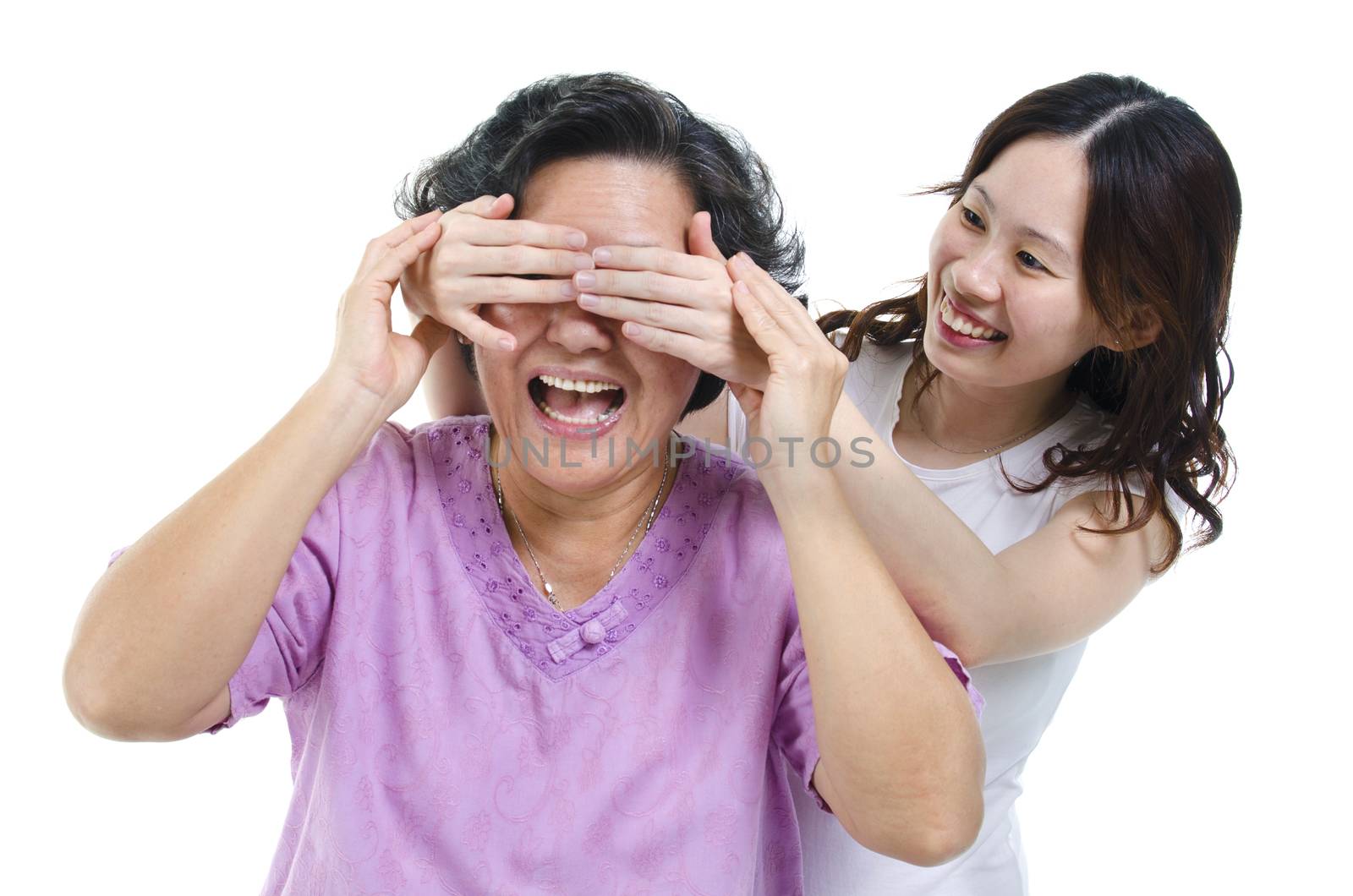 Portrait of Asian adult daughter covering her senior mother eyes, both laughing happily, isolated on white background.
