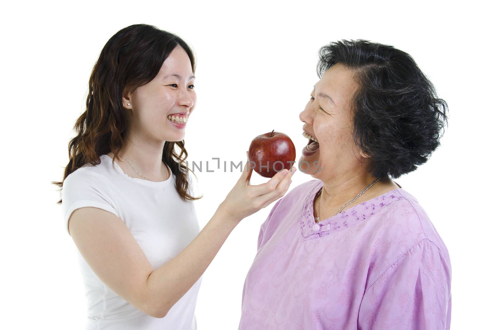 Portrait of Asian adult daughter feeding an apple to her senior mother and smiling, elderly healthy diet concept, isolated on white background.