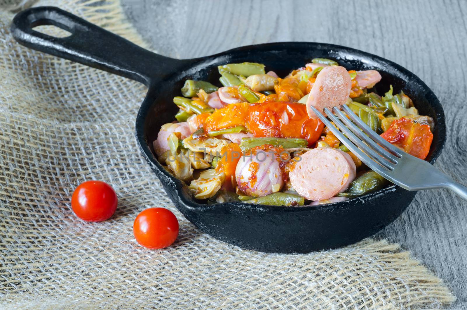Fried vegetables with chopped sausage in a cast iron pan, a piece on a fork. Gray wood background, burlap and selective focus.