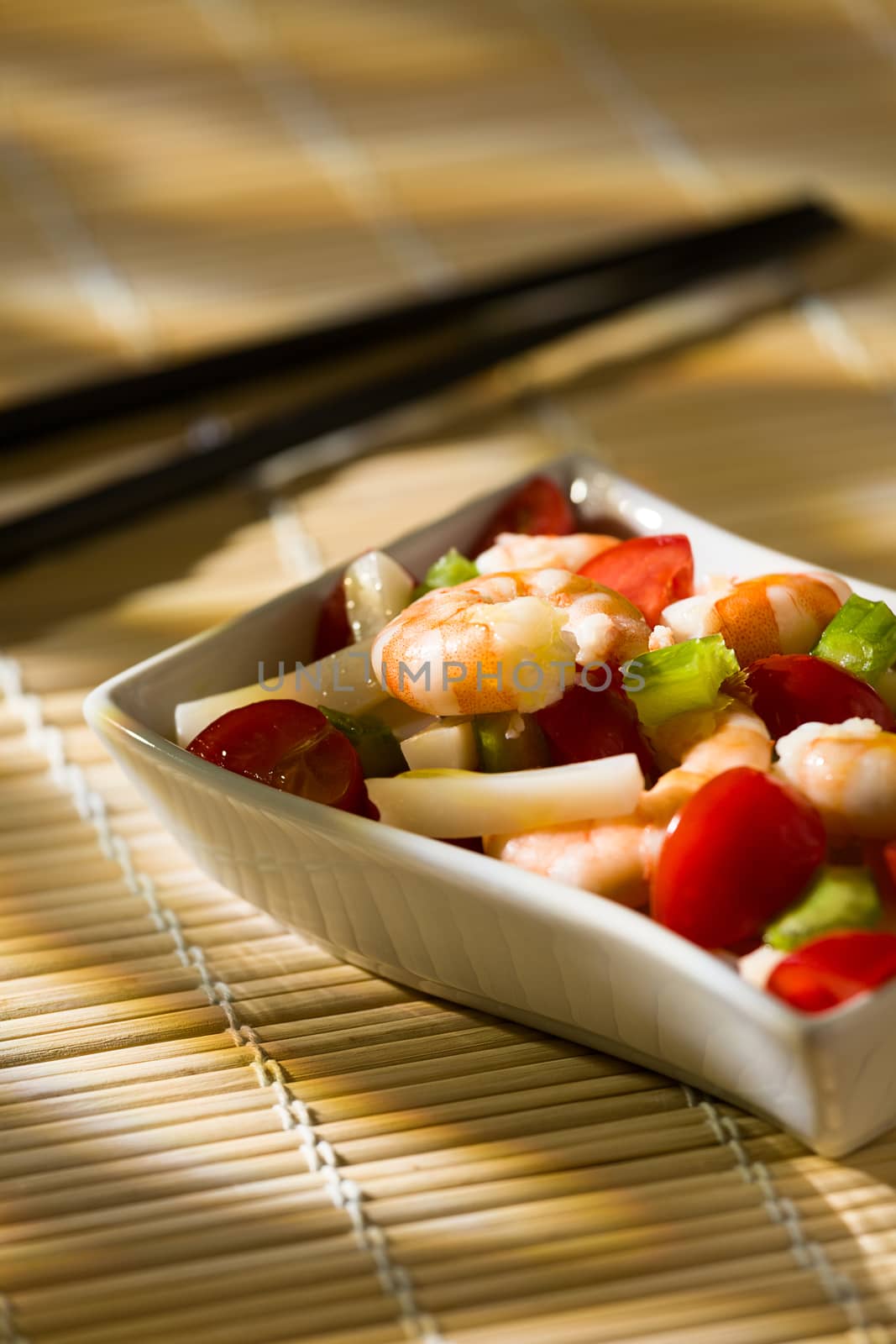 Shrimp salad with squid tomatoes celery inside a white bowl on a bamboo tablecloth and chopsticks