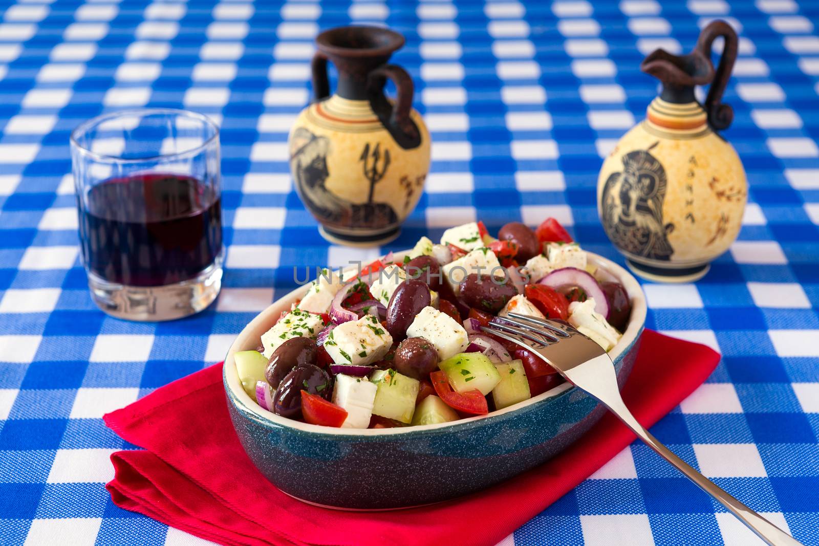 Greek salad with feta cheese tomatoes cucumber olives and onions with red wine and greek amphorae over a checkered tablecloth
