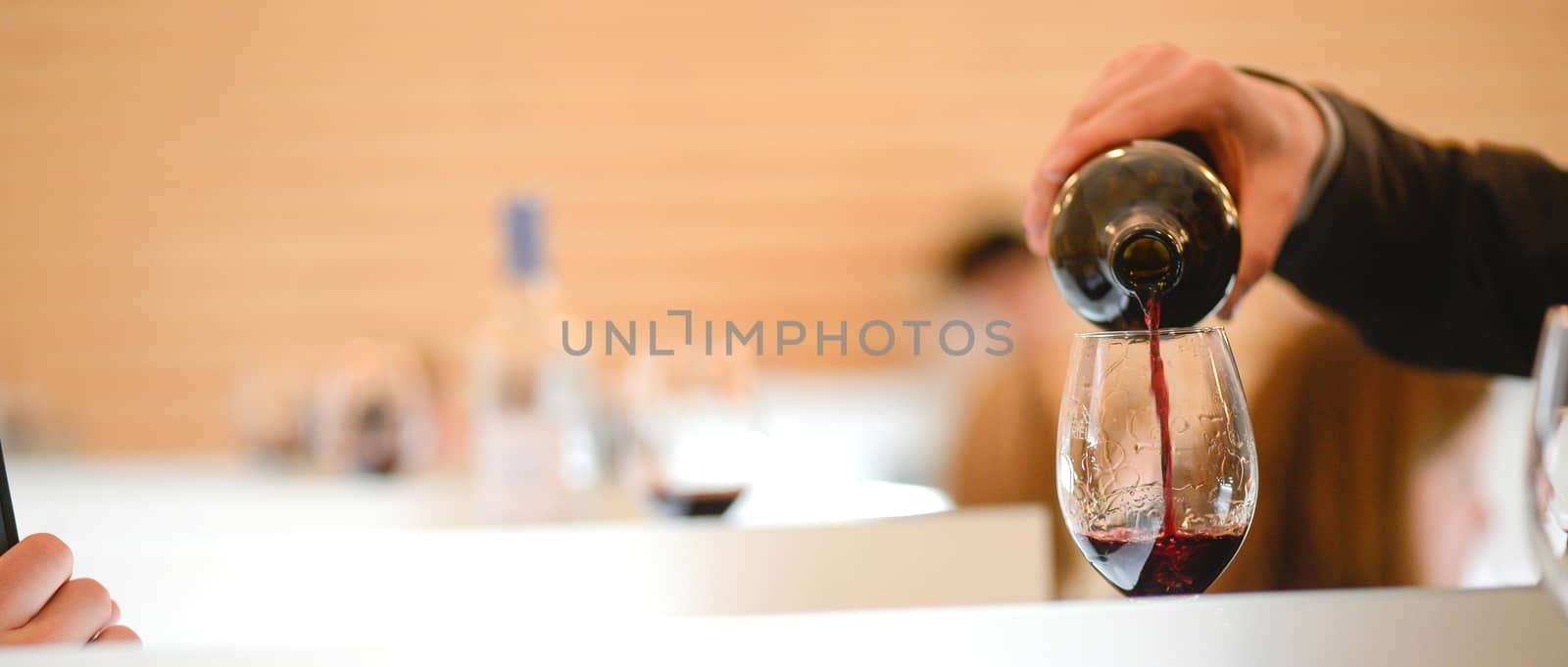 Tasting wine in a vinery-A man's hand is pouring red wine into a glass with a corkscrew set down. Lot of wine bottles in the background with shadows.