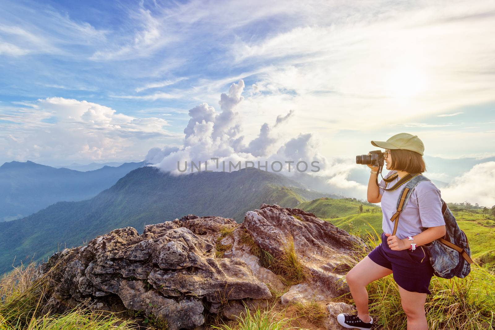 Hiker asian teen girl wear cap and eyeglass with happy holding dslr camera shooting photo beautiful nature in winter on peak mountain at sunset in Phu Chi Fa Forest Park, Chiang Rai, Thailand