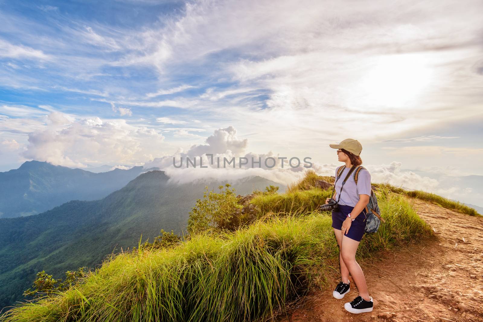 Hiker asian teen girl happy with camera backpack cap and eyeglass looking beautiful landscape nature of mountain range and colourful sky at sunset on Phu Chi Fa Forest Park, Chiang Rai, Thailand