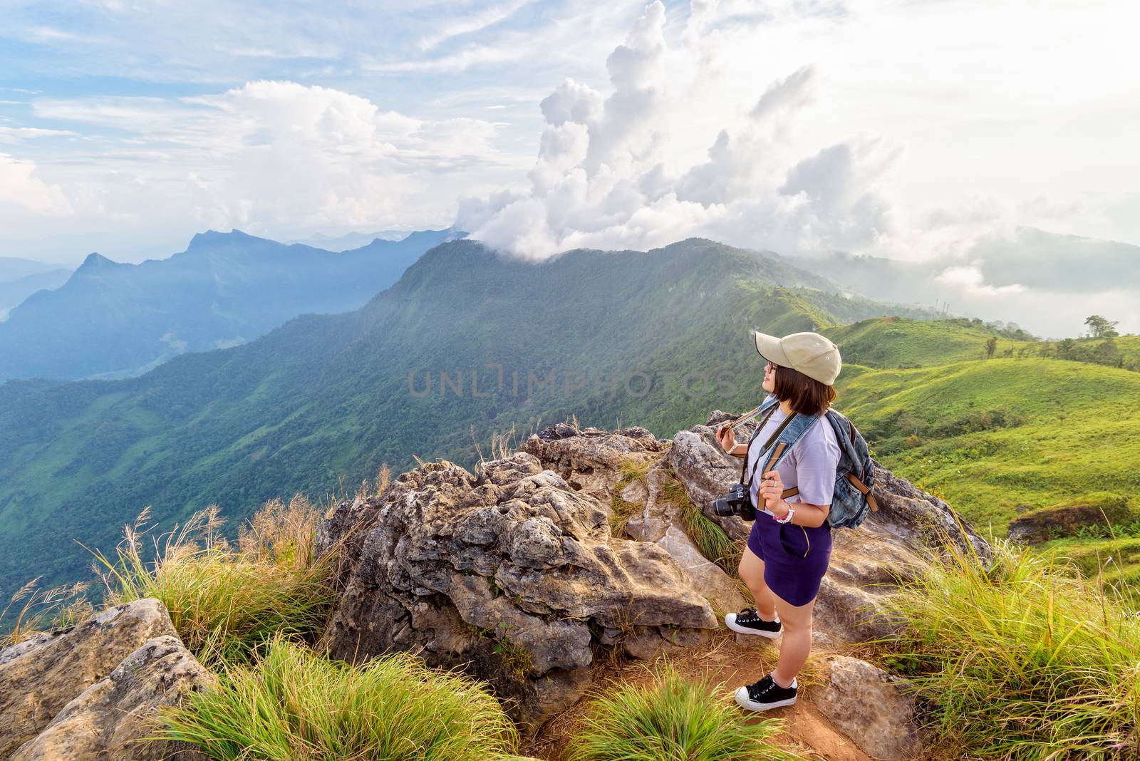 Hiker asian teen girl happy with camera backpack cap and eyeglass looking beautiful landscape nature of mountain range and colourful sky at sunset on Phu Chi Fa Forest Park, Chiang Rai, Thailand