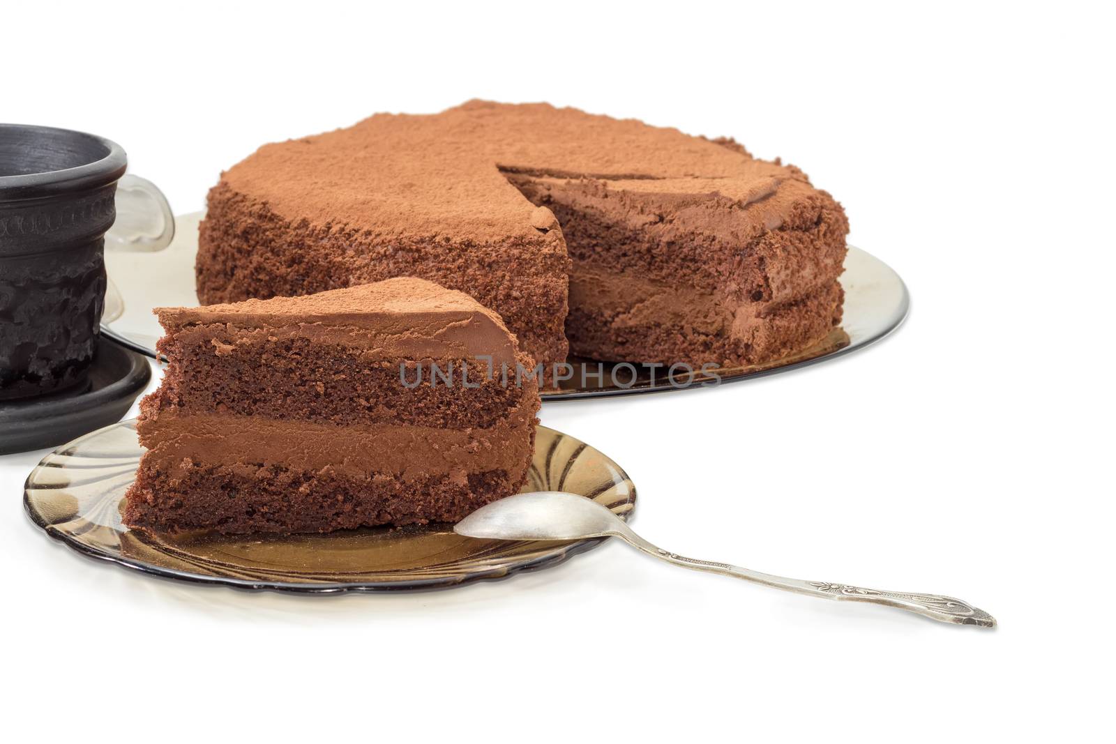 Piece of the chocolate layered cake sprinkled with cocoa powder and the spoon on a dark glass saucer on the background of the rest of the cake and coffee cup closeup on a light background
