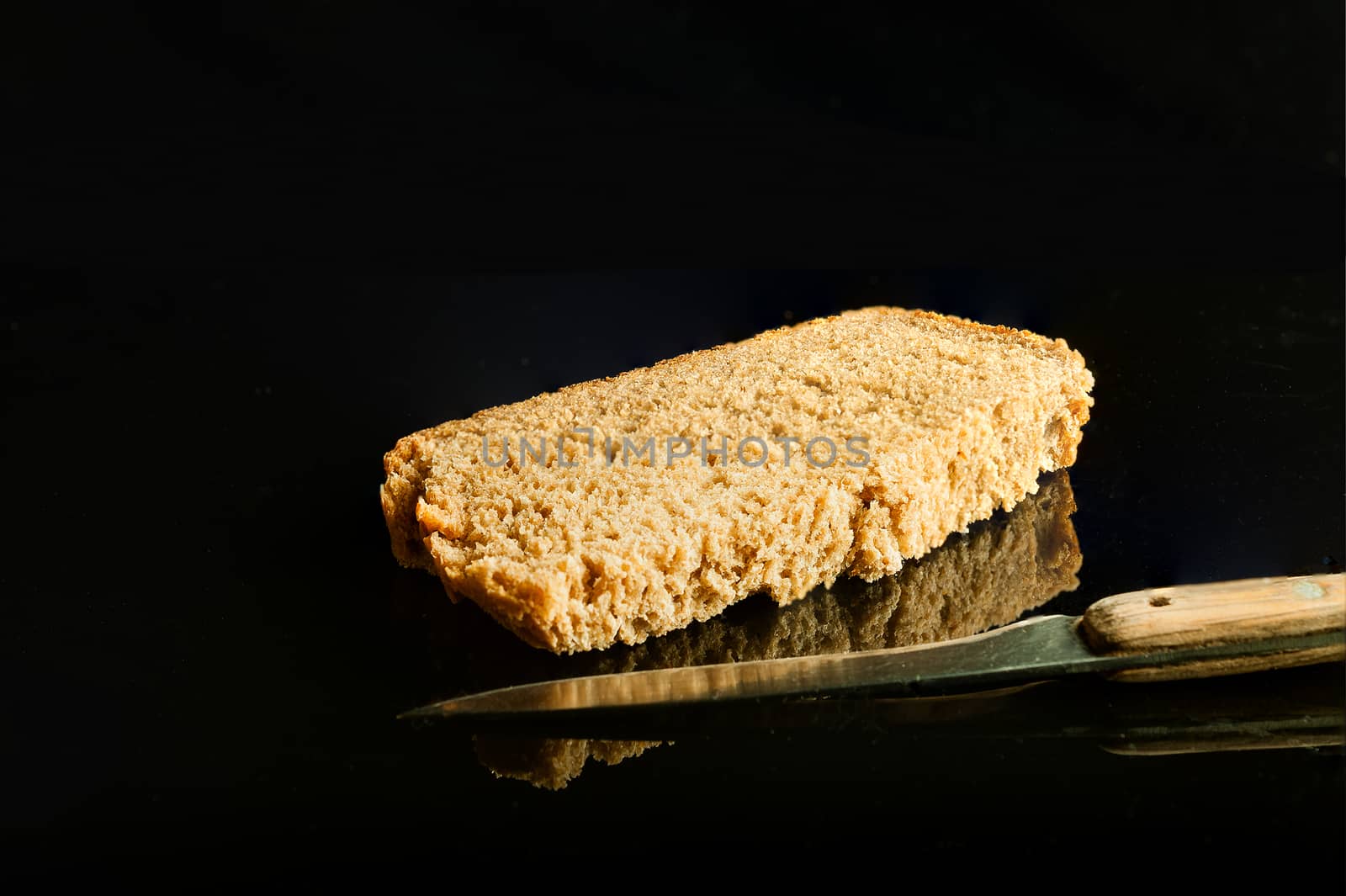 A slice of brown bread and a knife photographed in the studio on a black glass with reflection