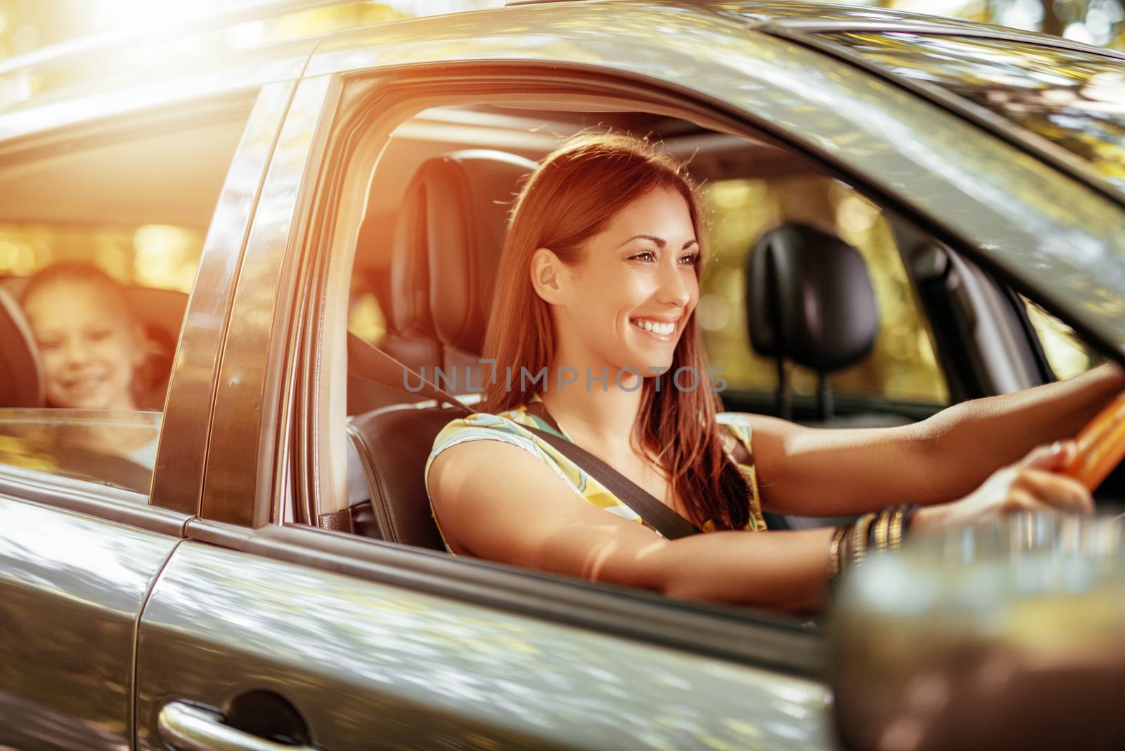 Young beautiful smiling woman driving a car.  Her cute daughter sitting on rear and enjoying.