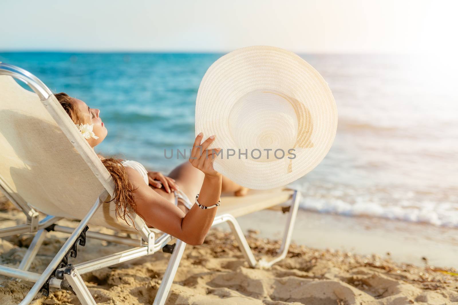 Beautiful young woman enjoying on the beach. She is lying on sunbed, holding summer hat and sunbathing. 