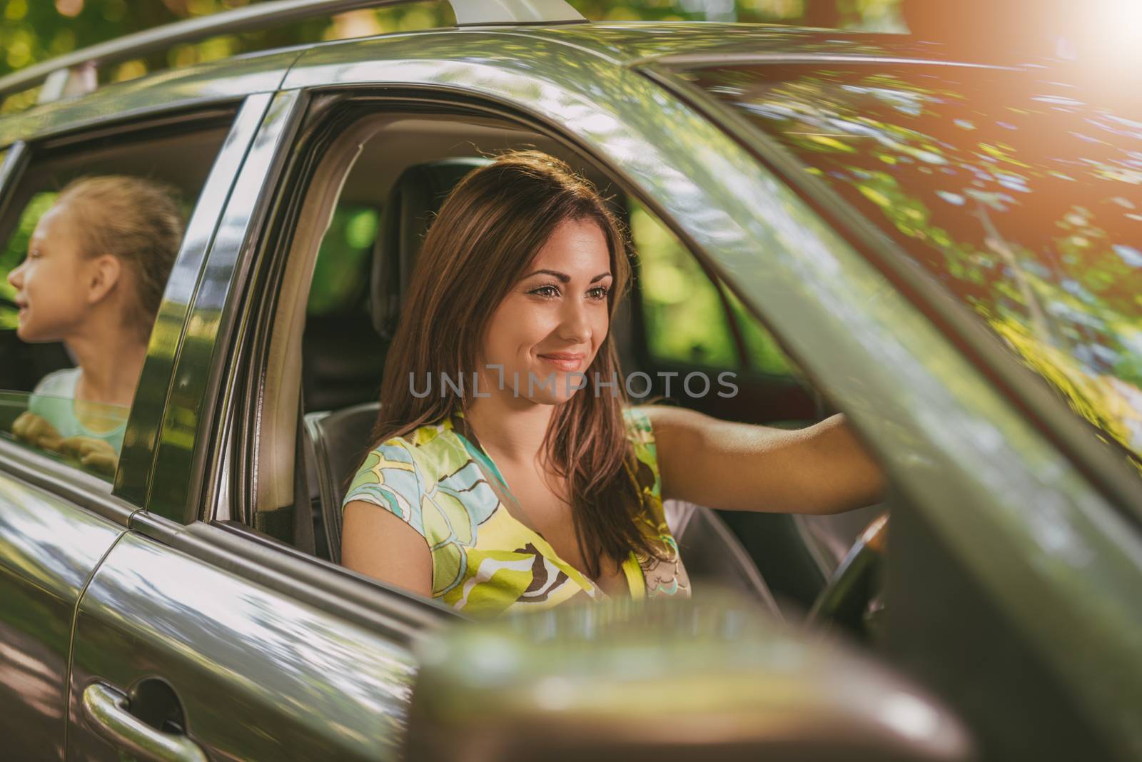 Young beautiful smiling woman driving a car.  Her cute daughter sitting on rear and looking the environment.