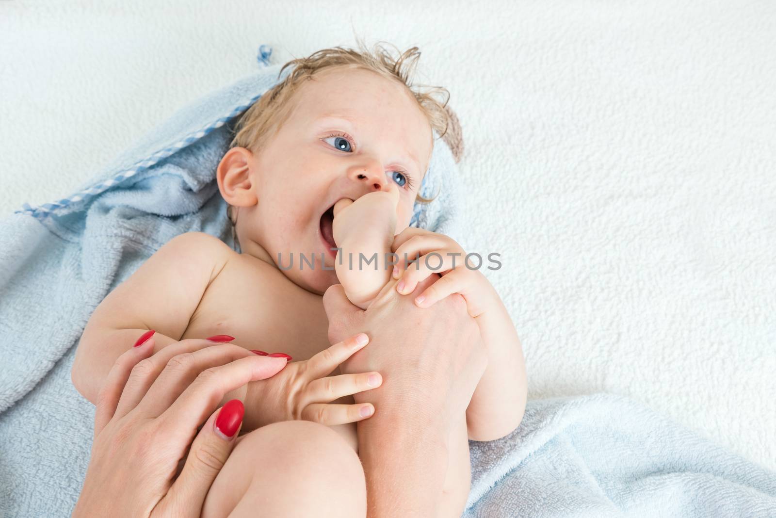 Cute happy beautiful infant boy playing with his foot with wet hair sitting in hothouse bath light blue fluffy towel white background, horizontal photo.