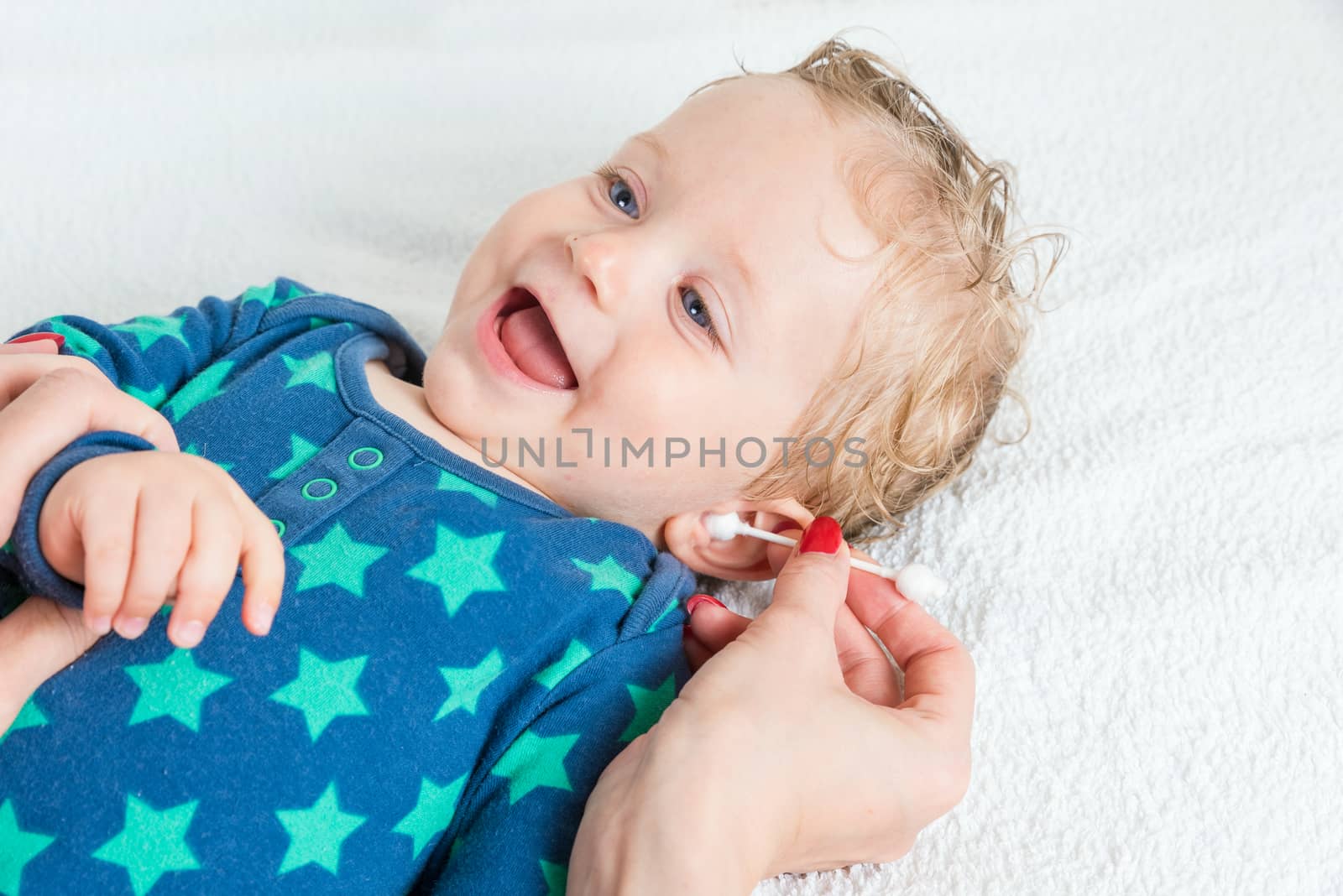 Mother hand cleaning baby ear with cotton swab,infant lying with wet hair and blue eyes and smiling,copy space,horizontal photo.