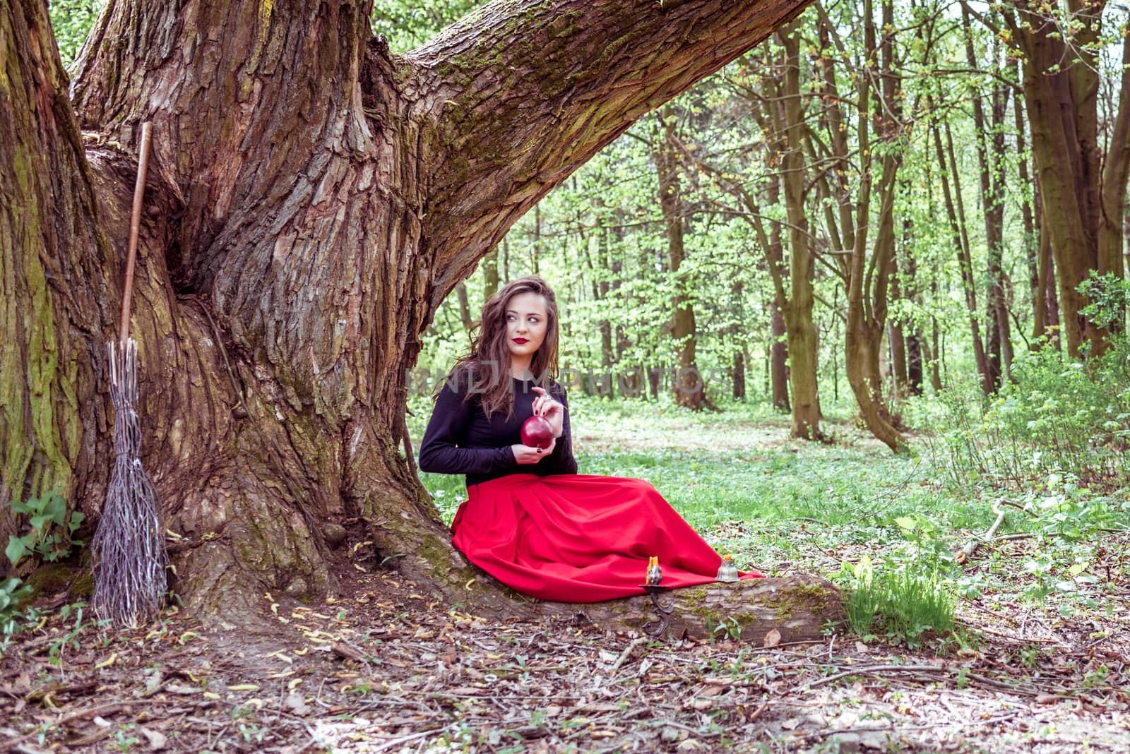 mystical witch woman in red dress sitting under a tree and holding an apple in the park